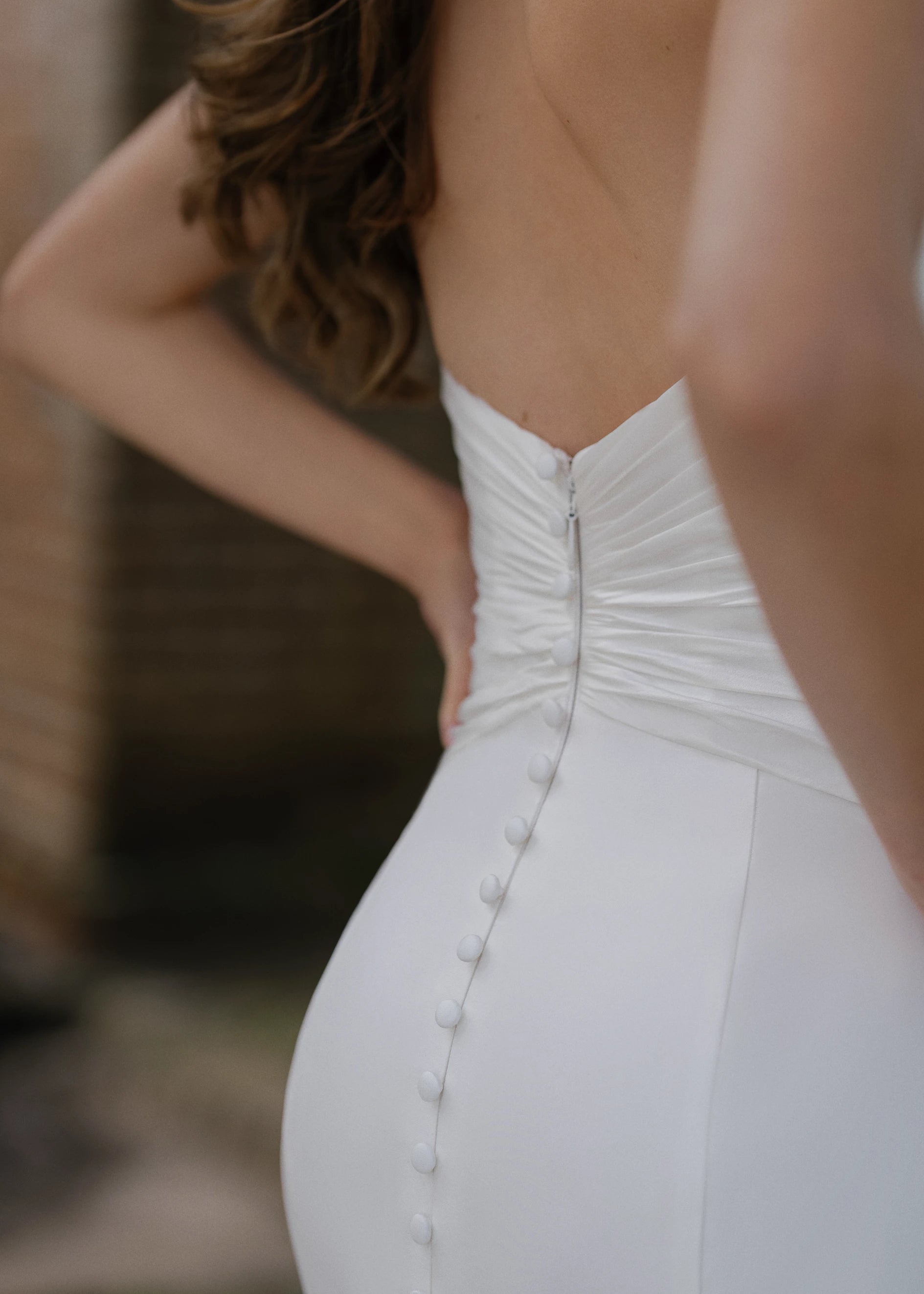 The Montagu gown is a trumpet gown of Italian Kalika with a narrow plunging V-neckline bodice, intricately pleated in Silk Satin Chiffon, and buttons to the top of the godet in Silk White.