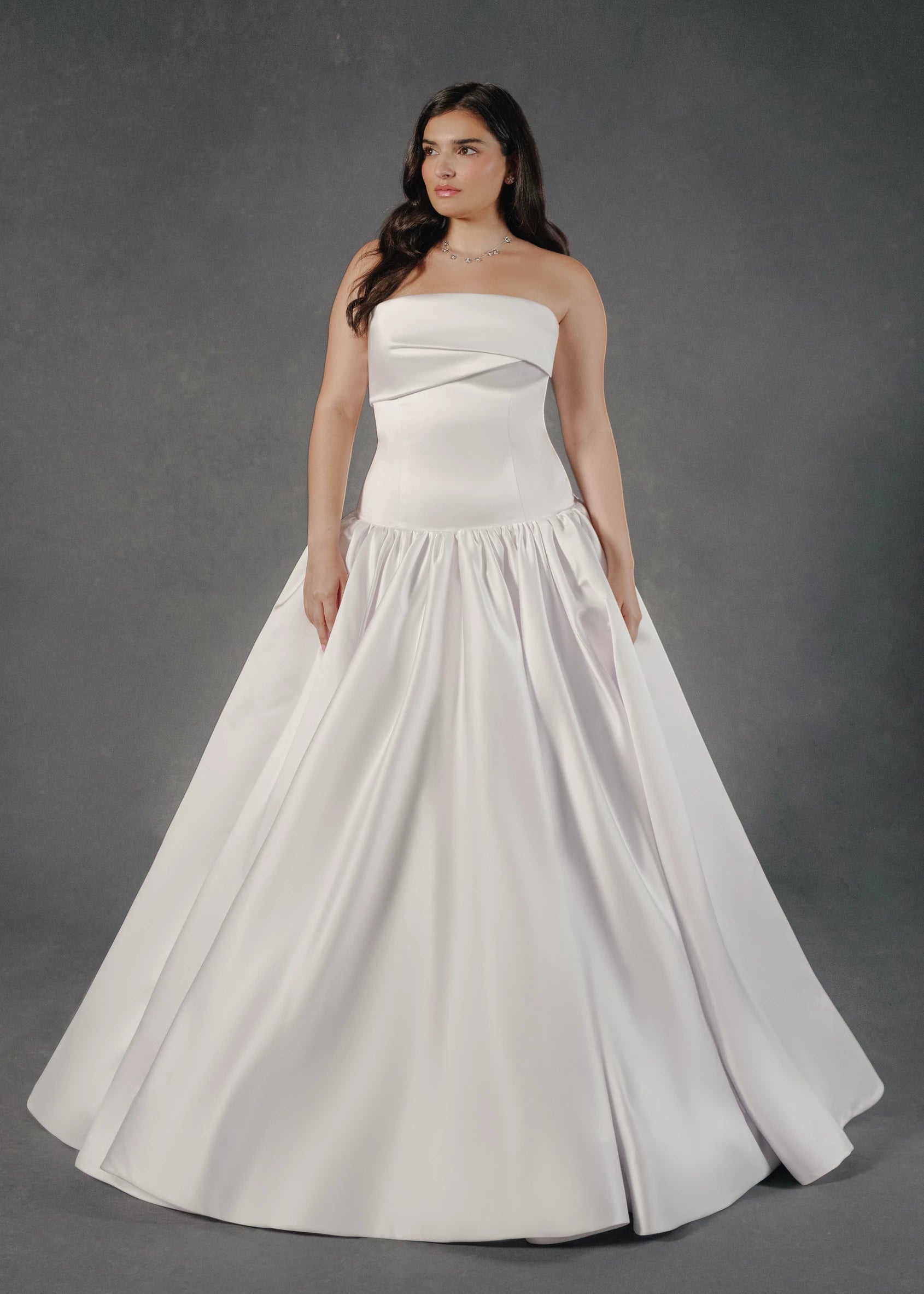 The Dakota wedding dress is a dramatic drop waist mermaid gown in Satin Back Faille with asymmetrically pleated neckline cuff, skirt gathers, and buttons to hem. 