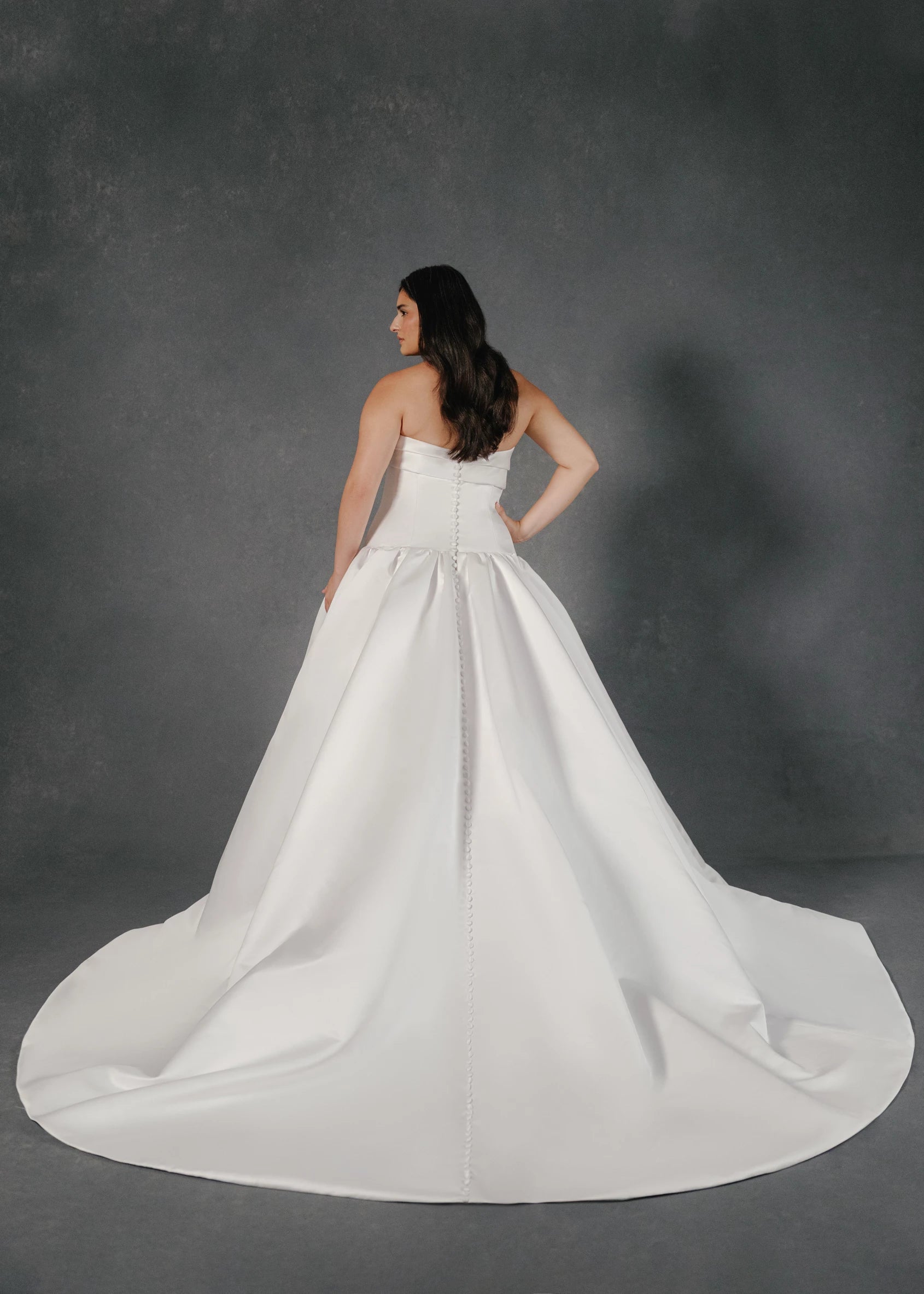 The Dakota wedding dress is a dramatic drop waist mermaid gown in Satin Back Faille with asymmetrically pleated neckline cuff, skirt gathers, and buttons to hem. 