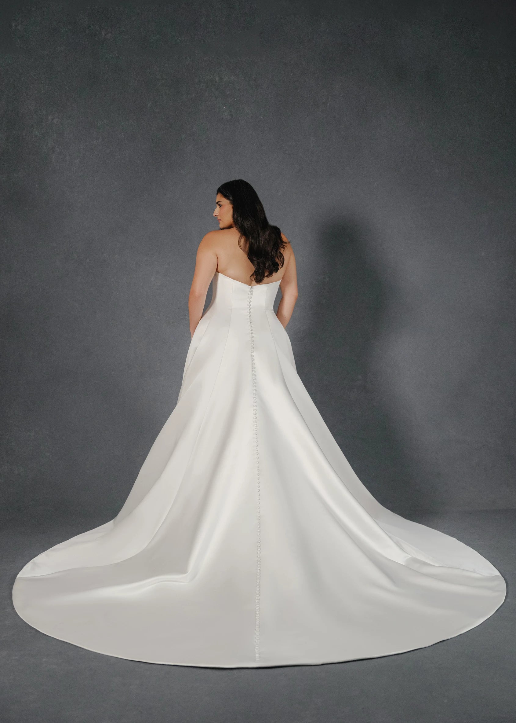 The Loyola gown is an arched strapless A-line gown of Faille-Back Satin with dramatic extended train and buttons to the hem in Silk White and Bright White. 