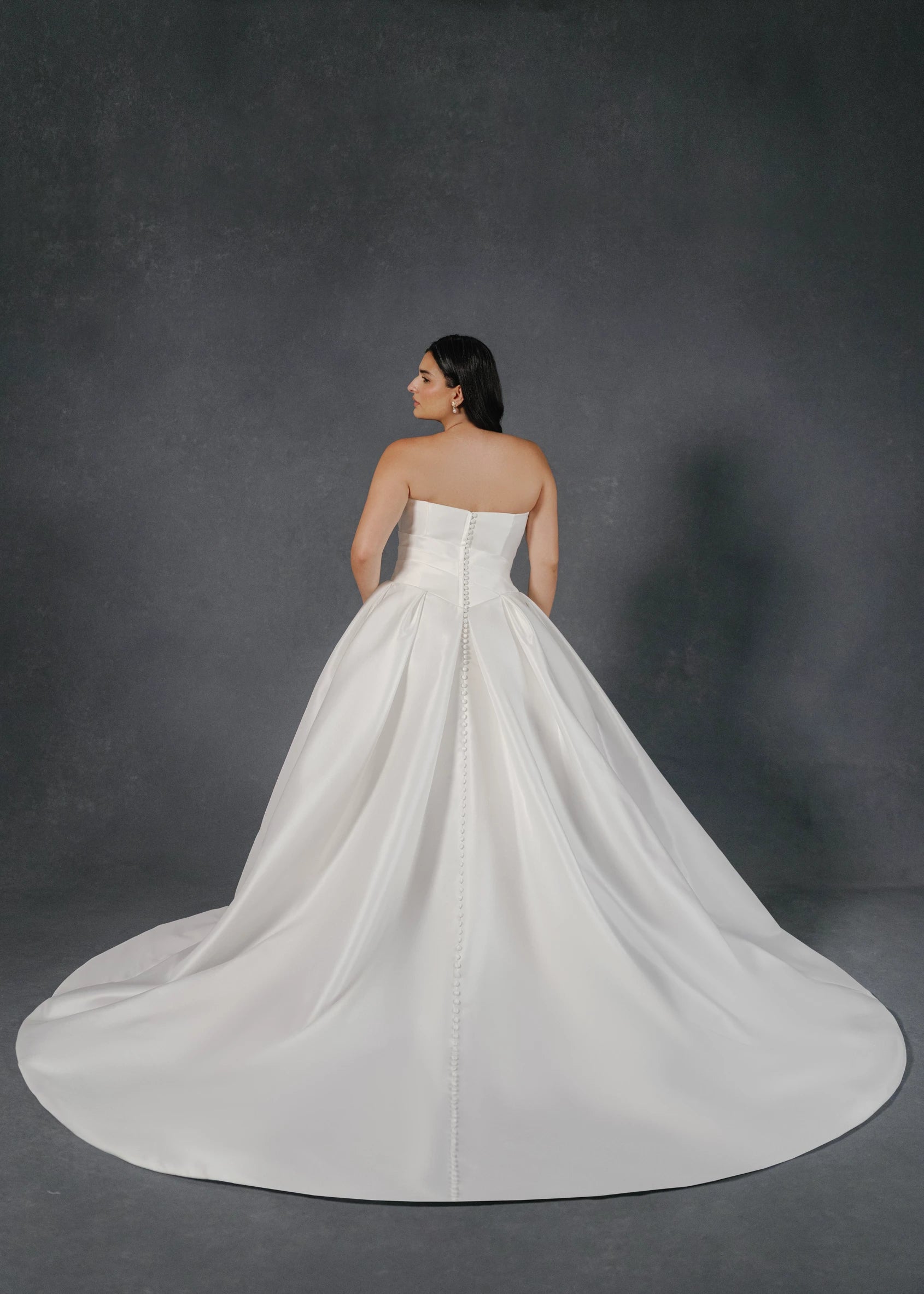 The Monarch gown is an arched strapless ballgown in Italian Lace Printed Matte Satin with asymmetrically draped bodice and basque waist on a dramatic box pleated skirt, with buttons to the hem in Pearl.