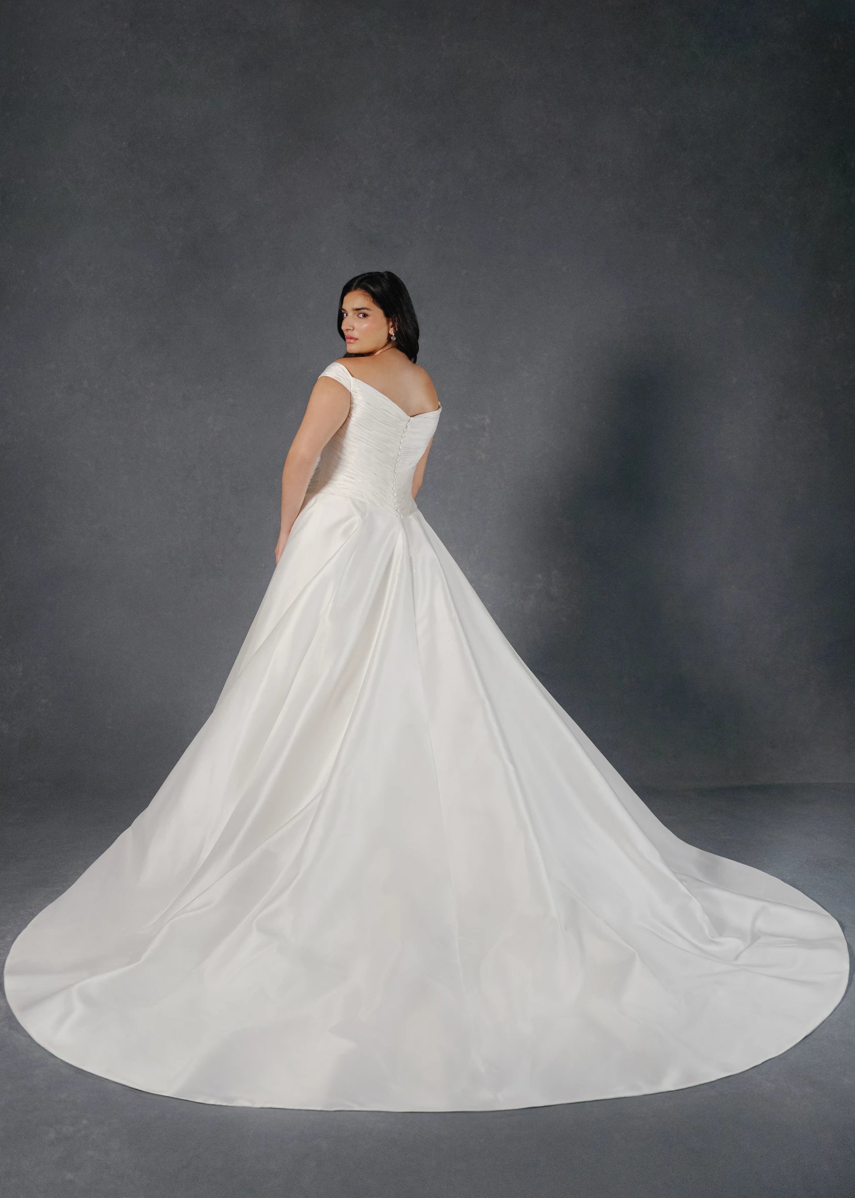 The Sabrina wedding dress is a timeless classic featuring a basque waist on a Silk White Satin Chiffon pleated, off-the-shoulder bodice with buttons to the waist and Kalika ballgown skirt.