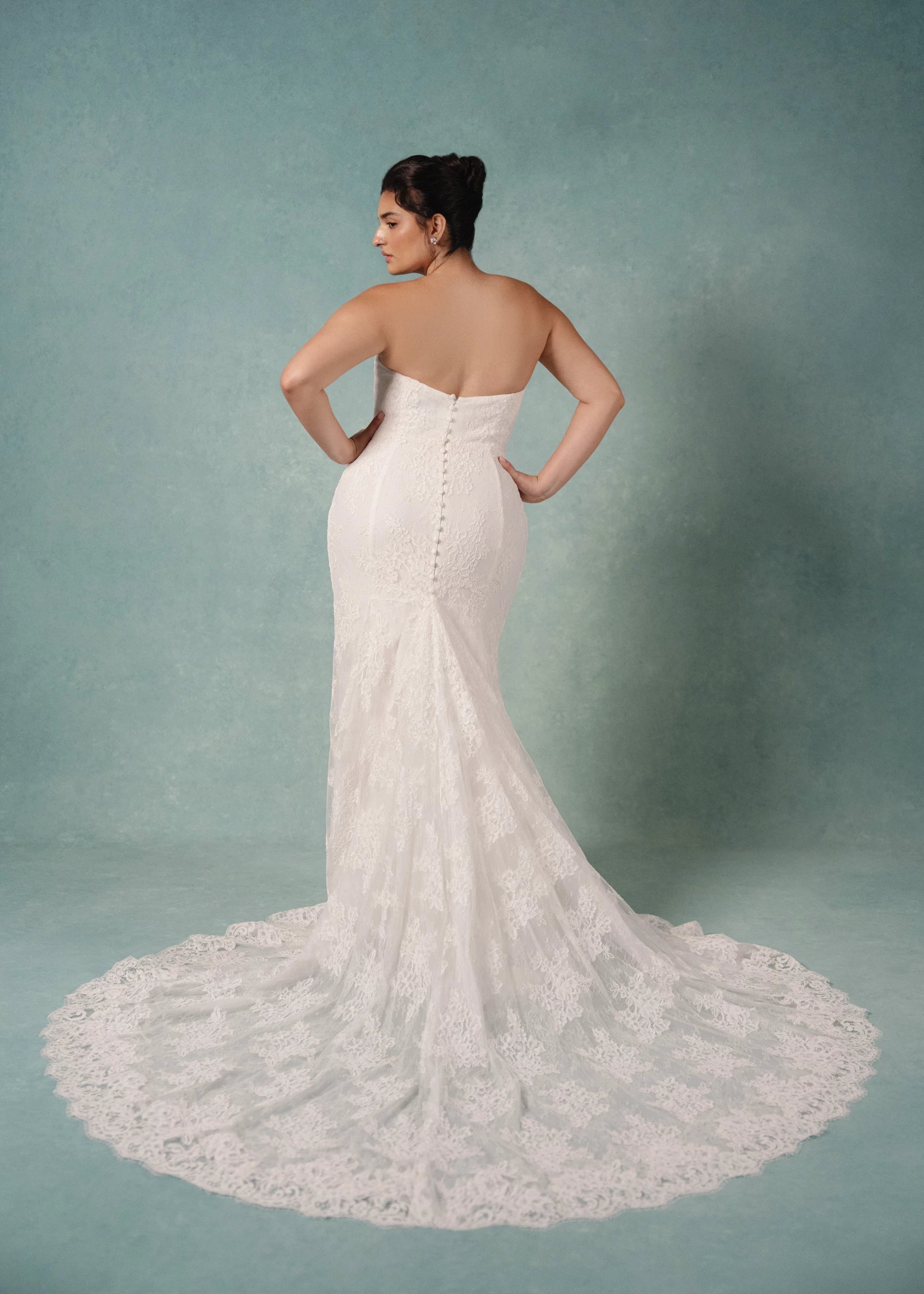 The Lucienne gown is a cat-eye strapless column gown of Corded Lace with pleated Illusion Tulle overlay, featuring buttons to the hem in Silk White.
