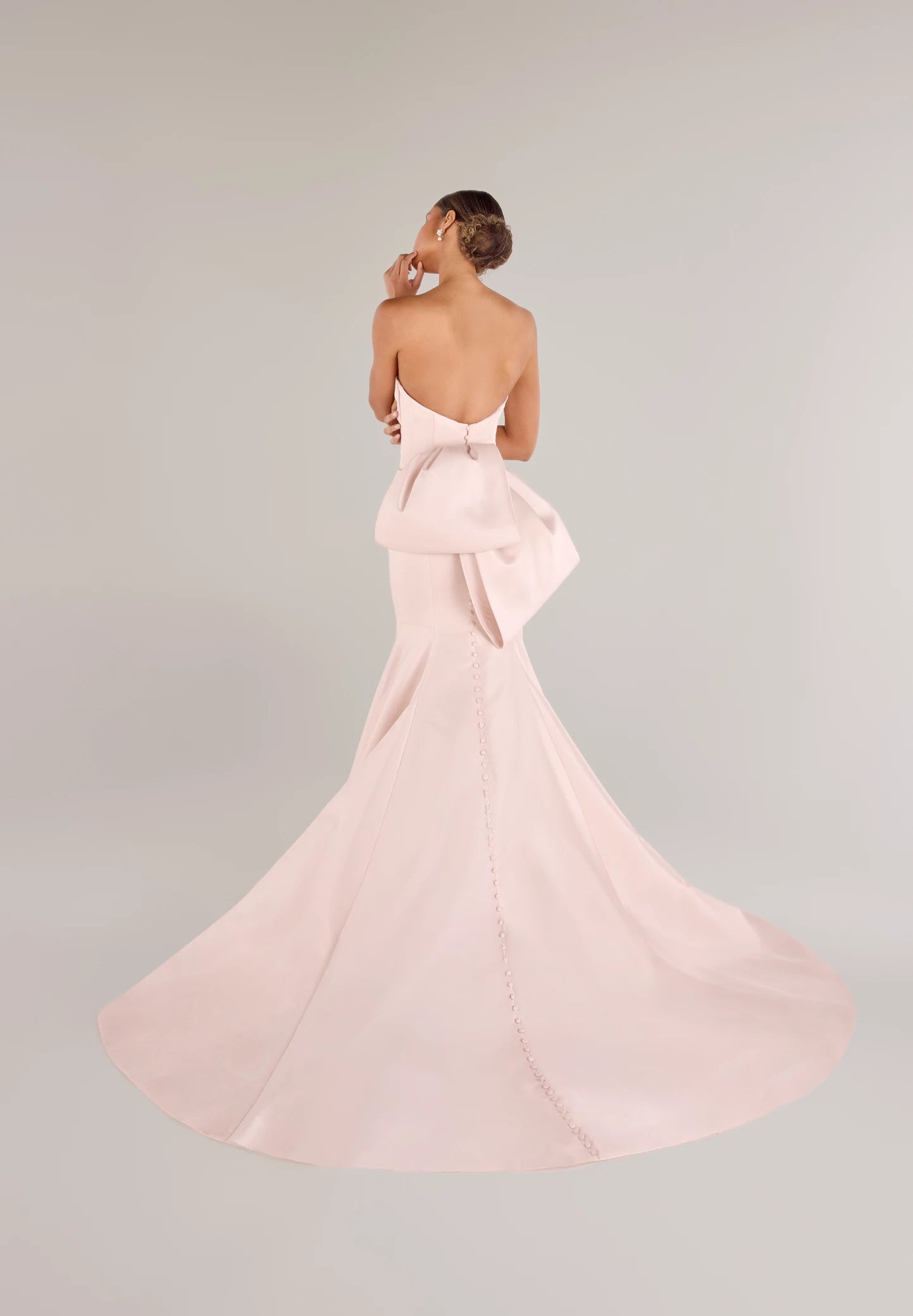 The Daniels gown is a fit and flare gown of Kalika featuring an asymmetrically draped bodice cuff, oversized sculptural bow, and buttons to the hem