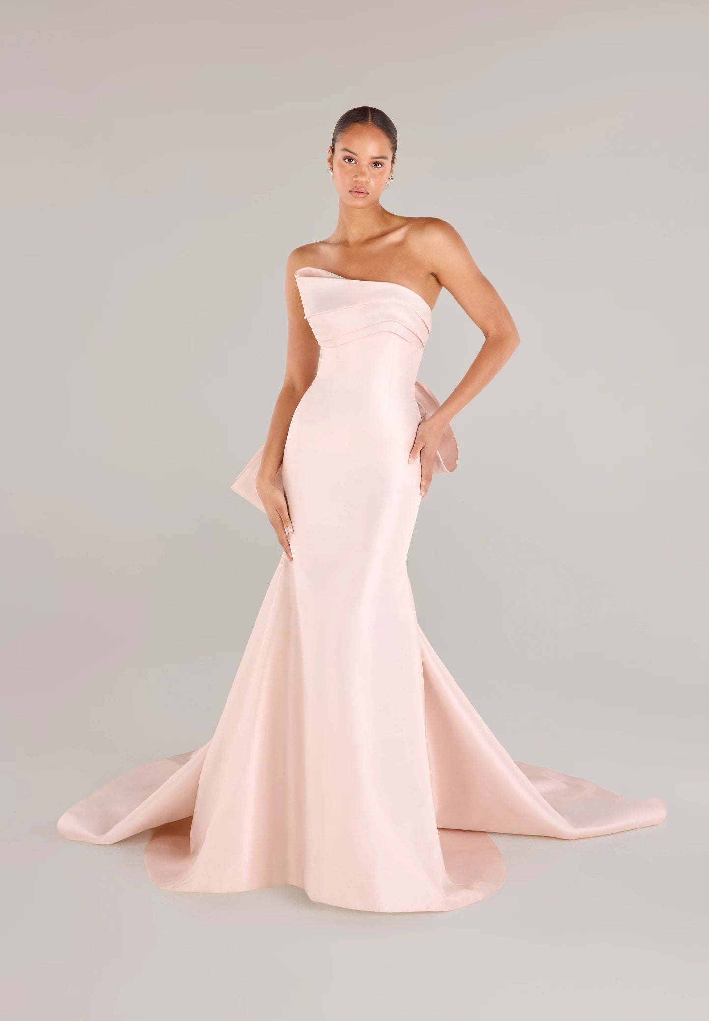 The Daniels gown is a fit and flare gown of Kalika featuring an asymmetrically draped bodice cuff, oversized sculptural bow, and buttons to the hem.