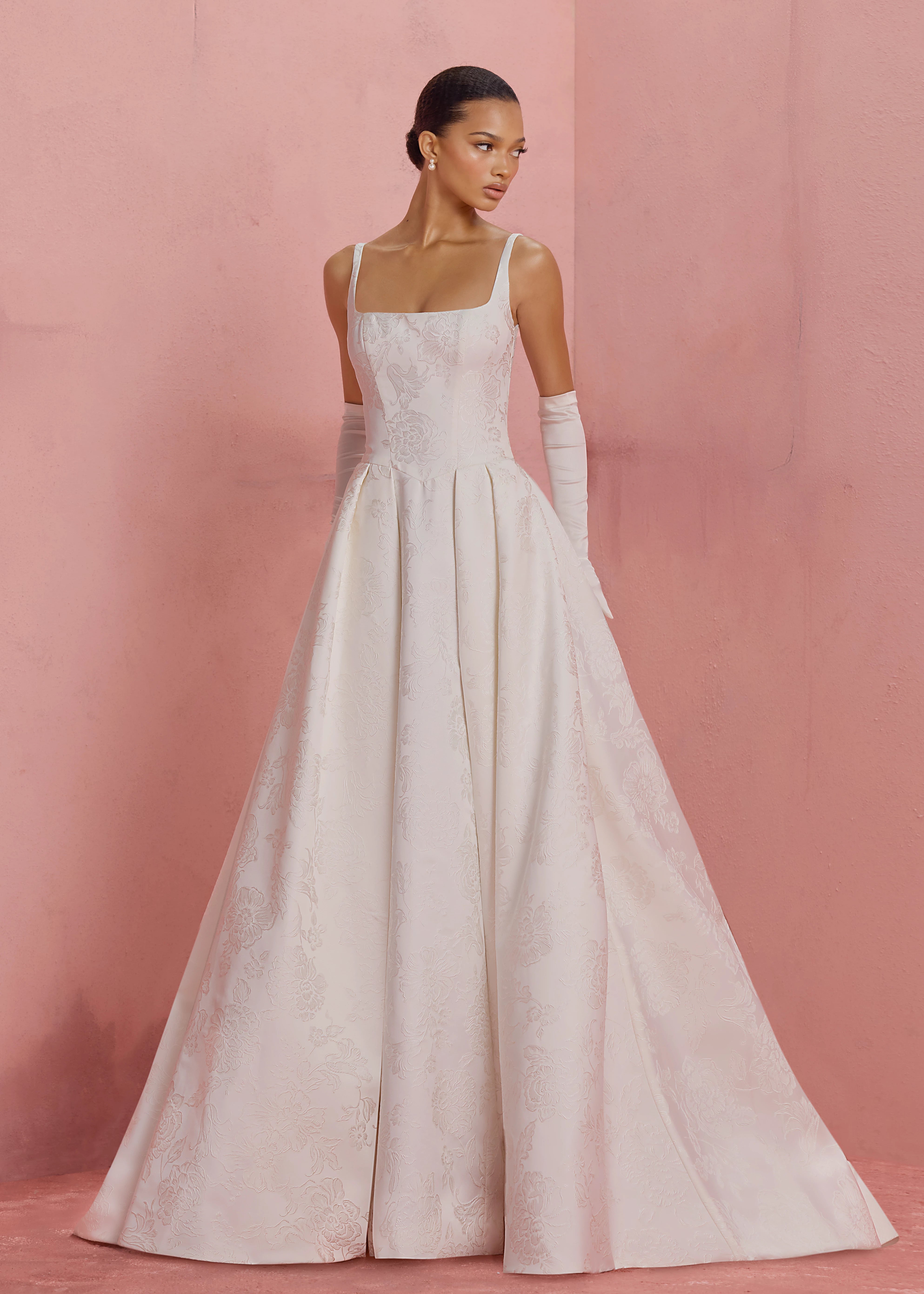 The Elodie gown by Anne Barge is a square neck column gown in Embossed Jacquard with low back and buttons to the hem, featuring box-pleated attached overskirt in Pearl.