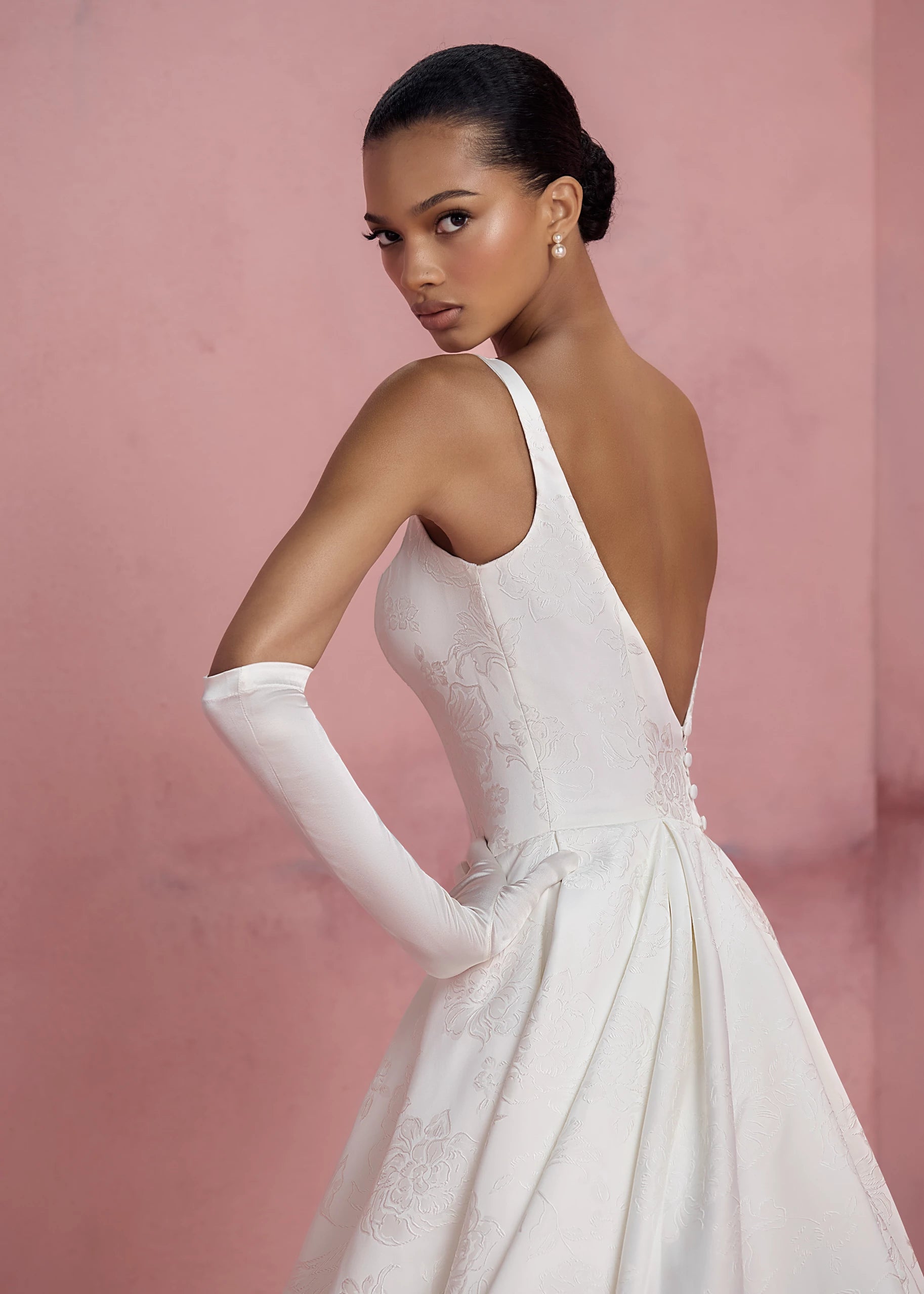 The Elodie gown by Anne Barge is a square neck column gown in Embossed Jacquard with low back and buttons to the hem, featuring box-pleated attached overskirt in Pearl.