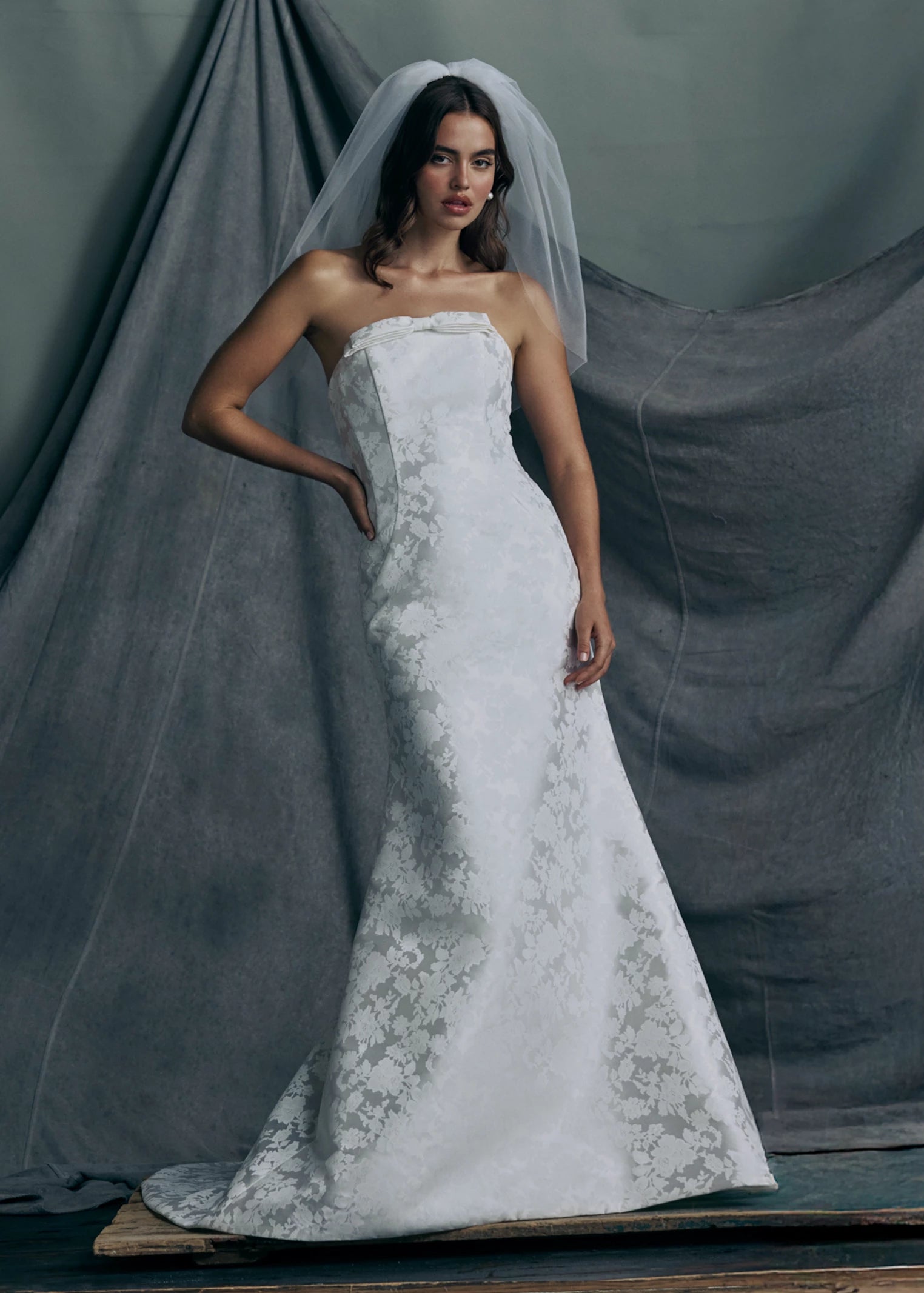 The Elysia gown by Anne Barge is an arched strapless gown in Italian Lace Printed Matte Satin with tailored bow detail, modified A-line skirt and buttons to the hem in Pearl.