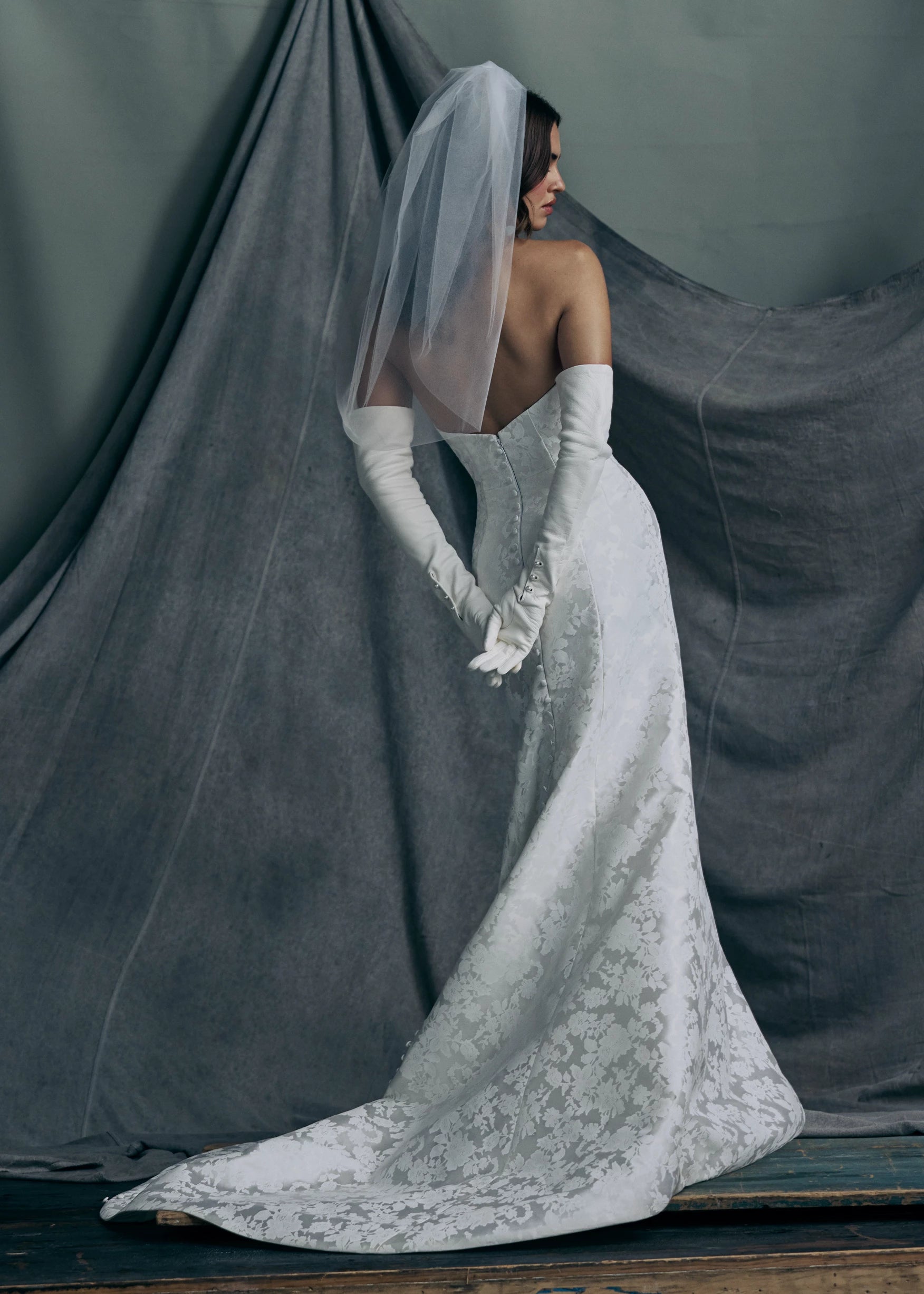 Back view of the Elysia gown by Anne Barge is an arched strapless gown in Italian Lace Printed Matte Satin with tailored bow detail, modified A-line skirt and buttons to the hem in Pearl.