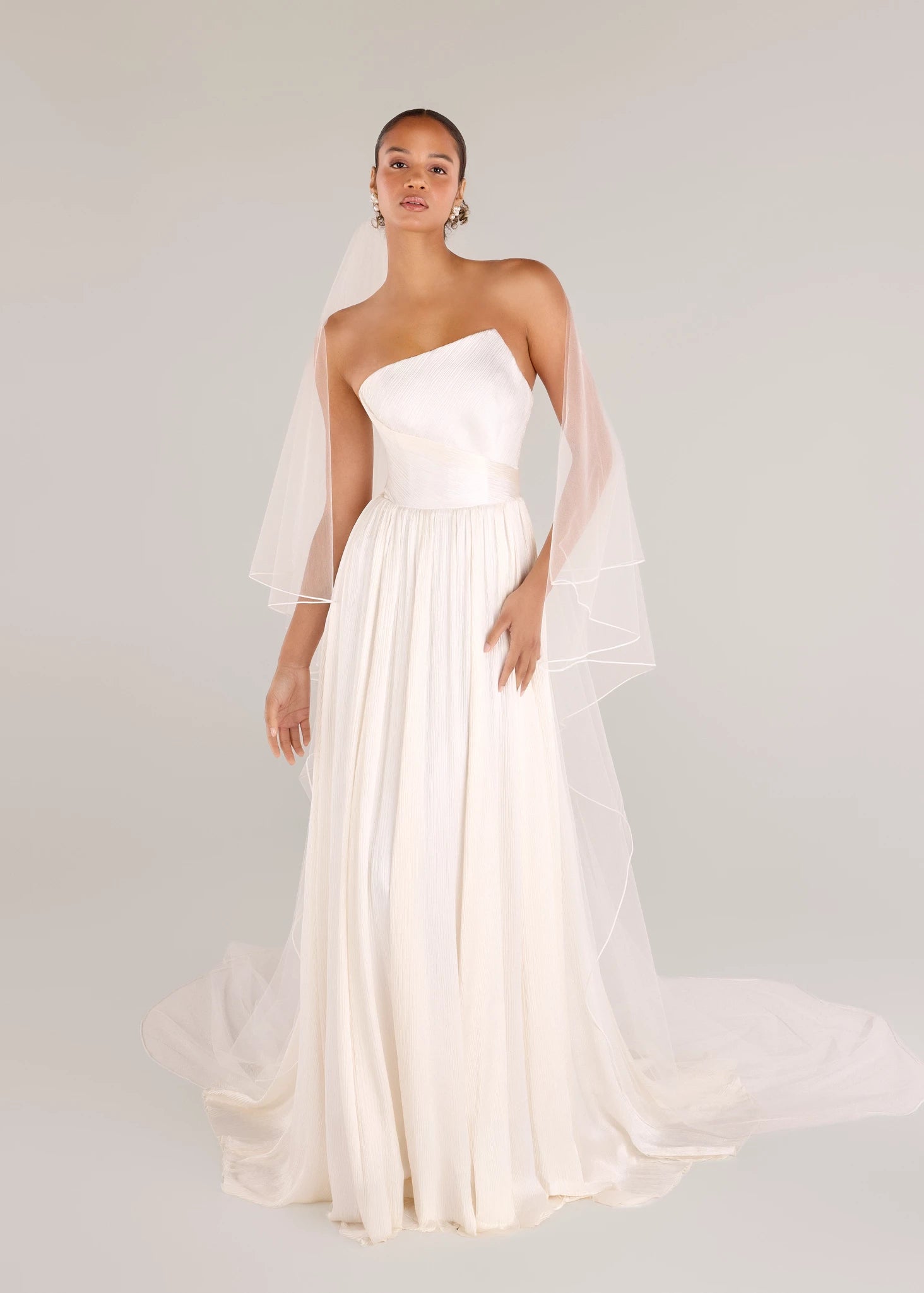 The Harelston gown is an A-line Crinkle Chiffon gown with softly gathered skirt and an asymmetrically pleated strapless bodice in Pearl.