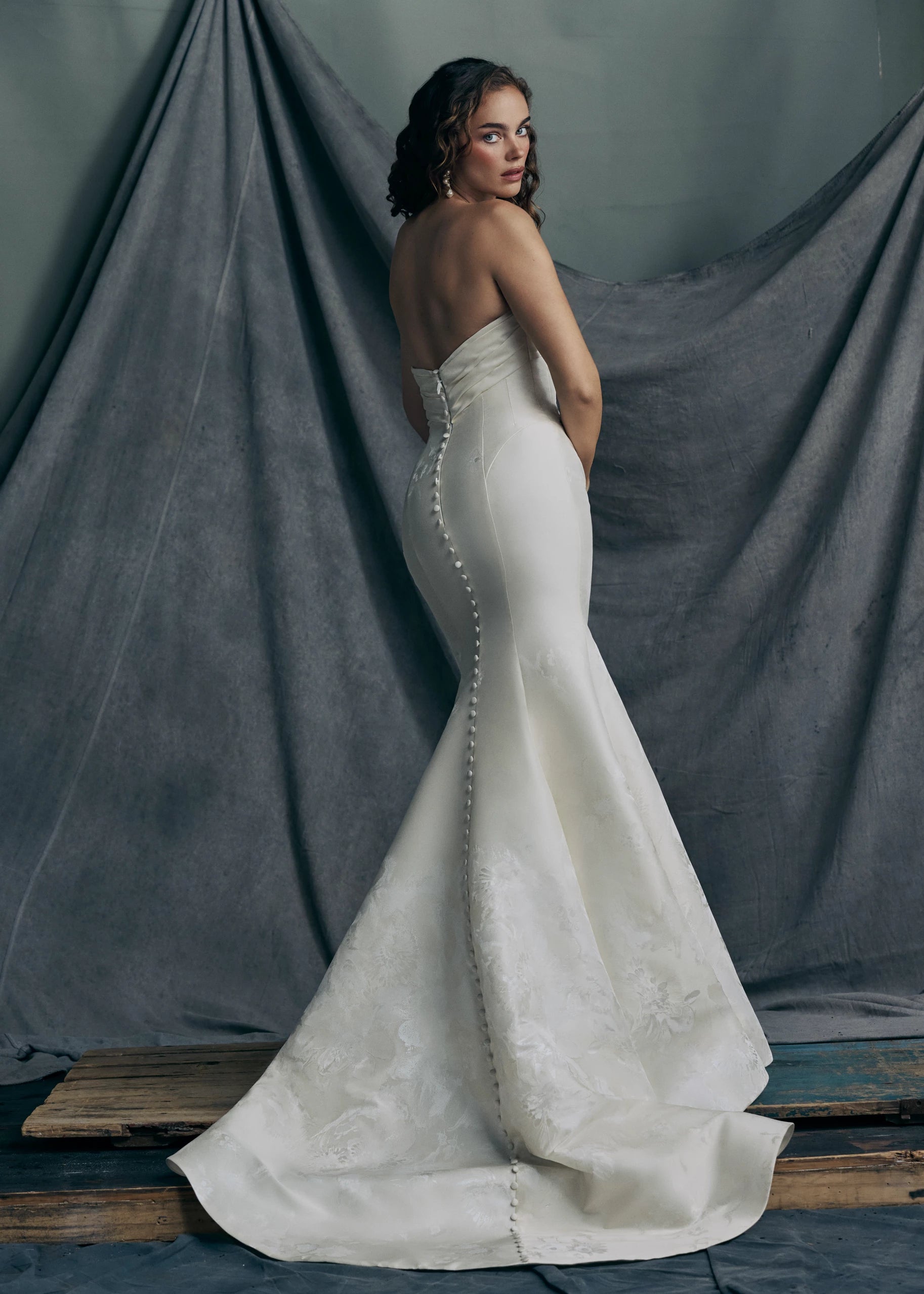 Back view of the Justine gown by Anne Barge is a strapless sweetheart trumpet gown of Italian Floral Matte Satin Jacquard with twisted draped bodice and buttons to the hem in Pearl.