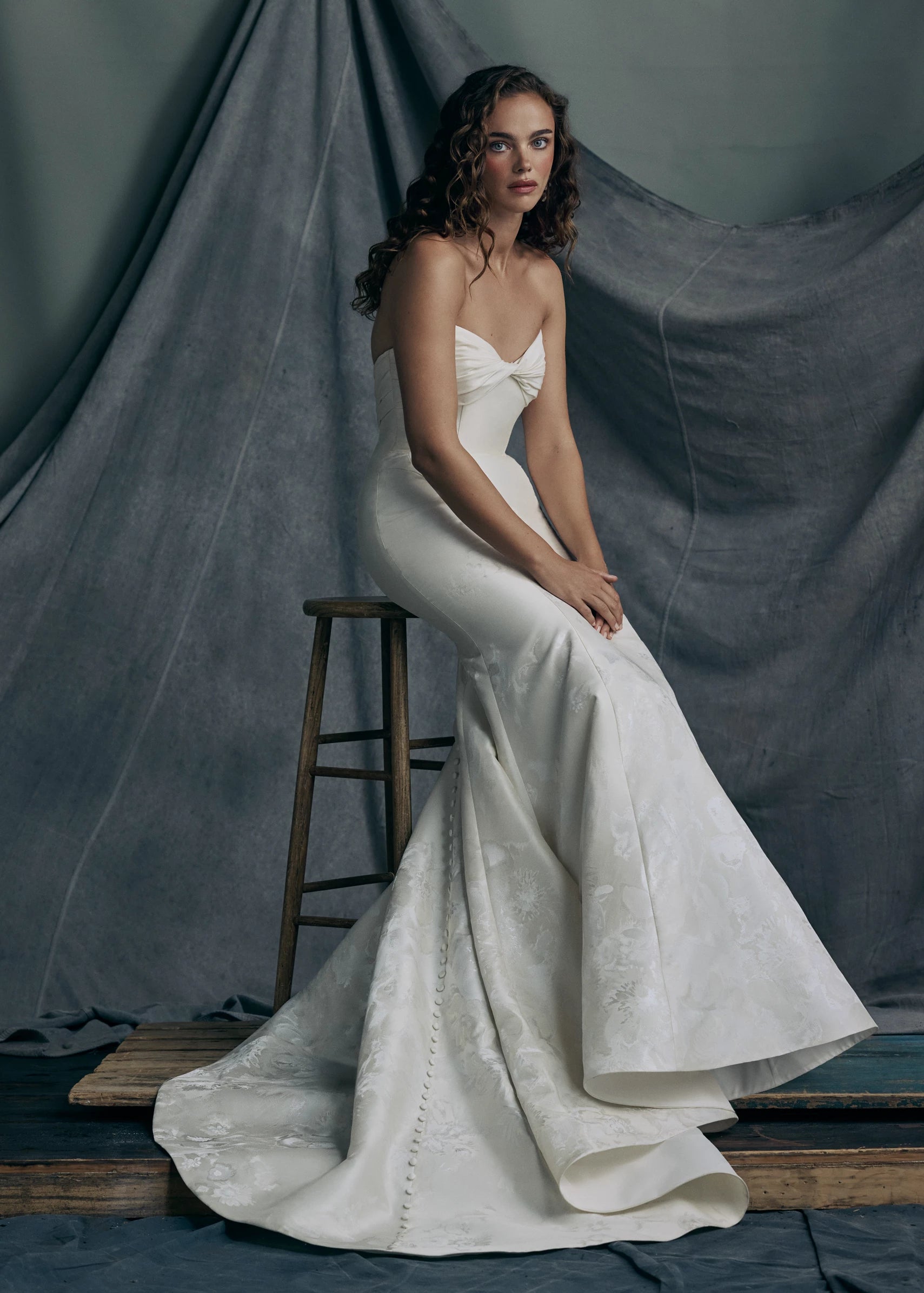 The Justine gown by Anne Barge is a strapless sweetheart trumpet gown of Italian Floral Matte Satin Jacquard with twisted draped bodice and buttons to the hem in Pearl.