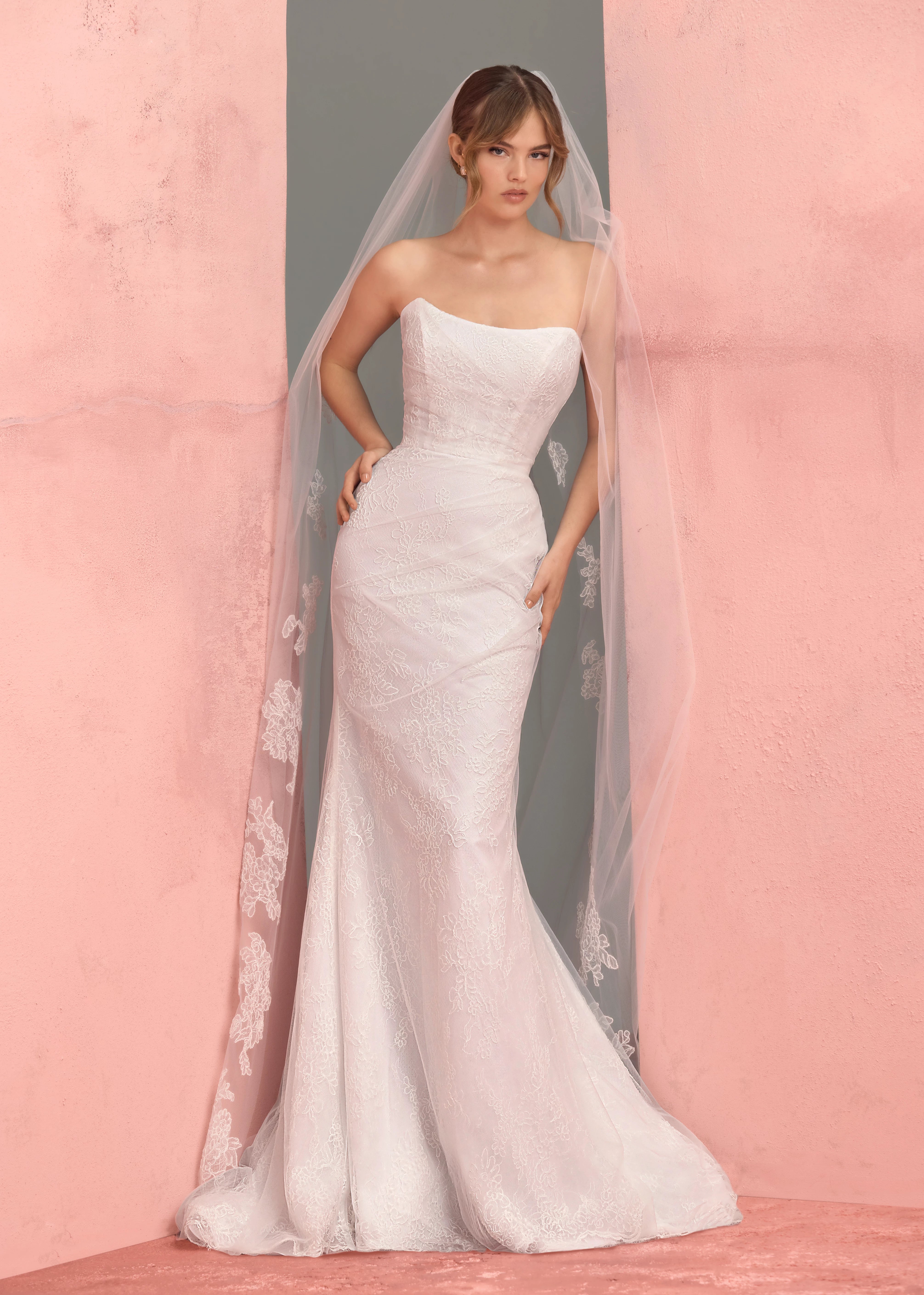 The Lucienne gown by Anne Barge is a cat-eye strapless column gown of Corded Lace with pleated Illusion Tulle overlay, featuring buttons to the hem in Silk White.