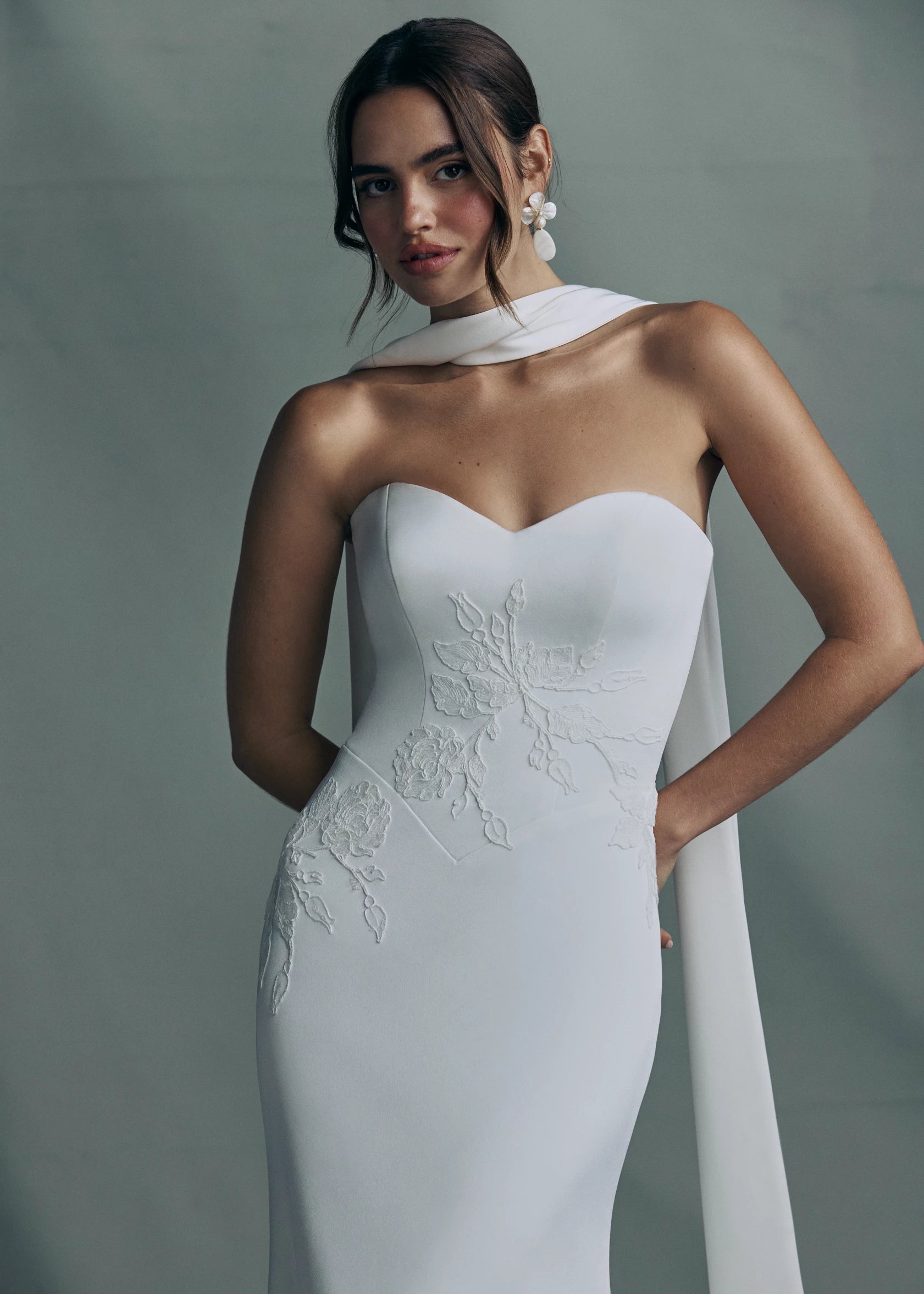 The Luminaire gown by Anne Barge is a strapless sweetheart gown in Crepe Back Satin, accented with Vine Embroidered Lace appliqués and pointed basque waist seam, with buttons to the hem in Silk White. Shown with the Luminaire Scarf, a neck scarf of Crepe Back Satin with Vine Embroidered Lace appliques at the edges.