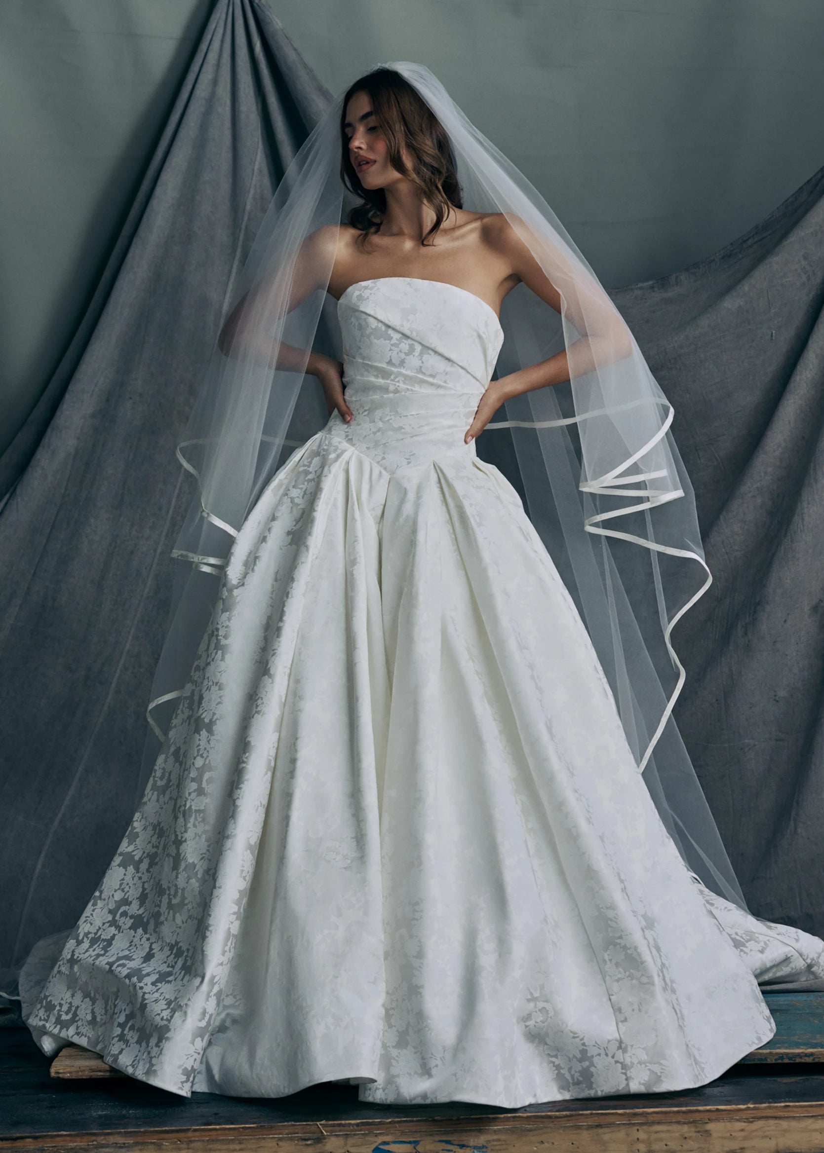 The Monarch gown by Anne Barge is an arched strapless ballgown in Italian Lace Printed Matte Satin with asymmetrically draped bodice and basque waist on a dramatic box pleated skirt, with buttons to the hem in Pearl.