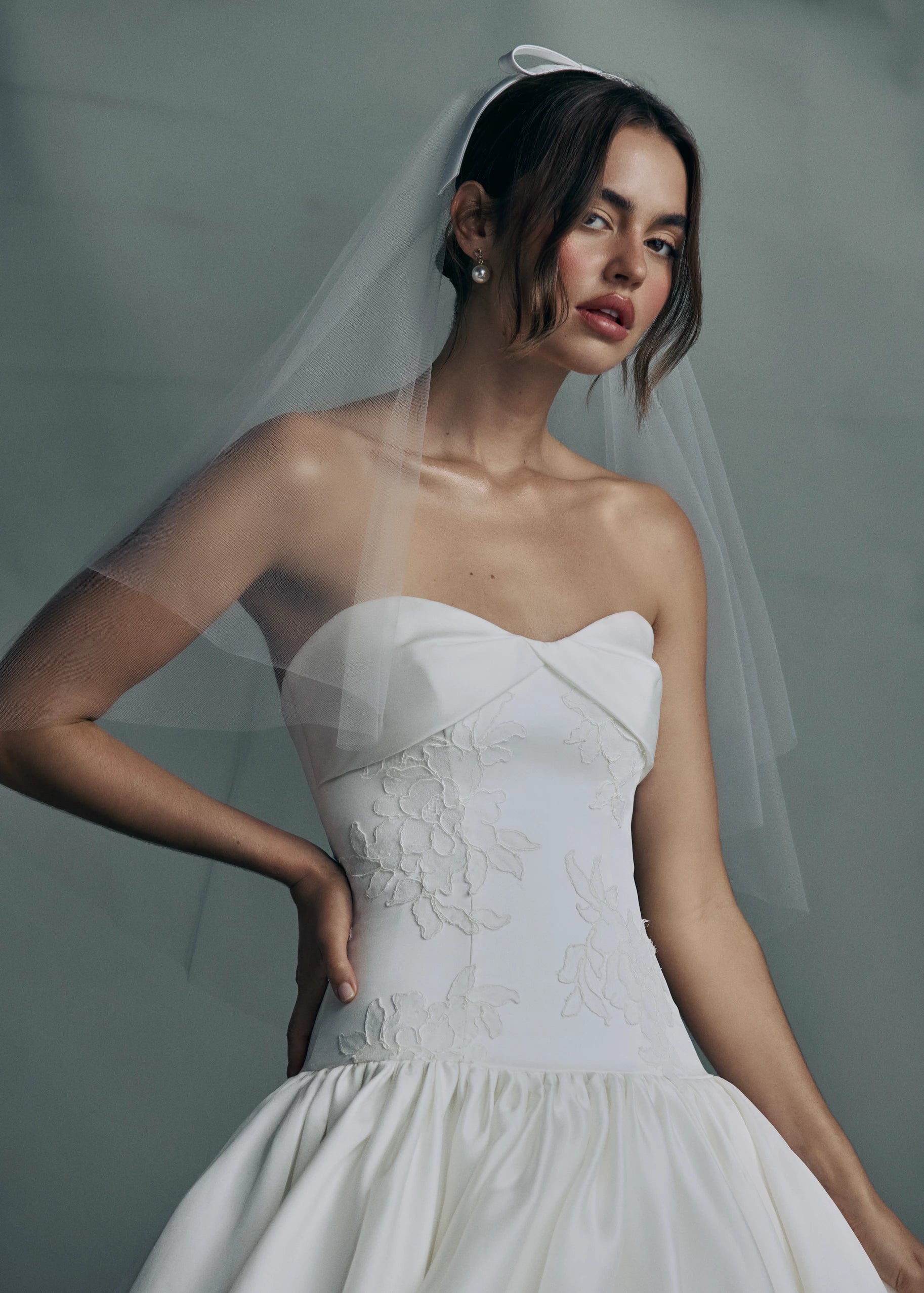The Noblesse gown by Anne Barge is a contour strapless drop waist ball gown of Silk Satin Organza with a draped neckline, delicate French Alençon lace appliqués, and a gathered skirt finished with buttons to the hem in Silk White.