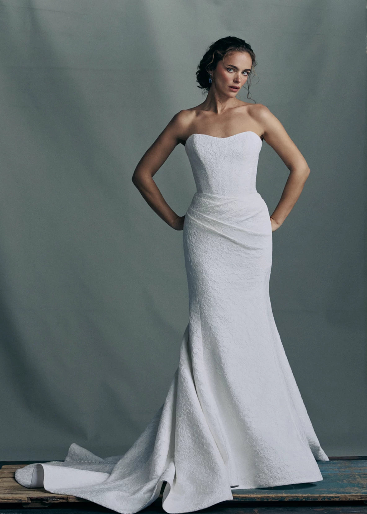 The Sera gown by Anne Barge is a strapless rounded cat-eye fit and flare gown in Spanish Lacework Jacquard, accented by diagonal draping across the skirt and buttons to the hem in Silk White. The Sera gown includes the Sera Shawl, a detachable off-the-shoulder shawl in Lacework Jacquard.