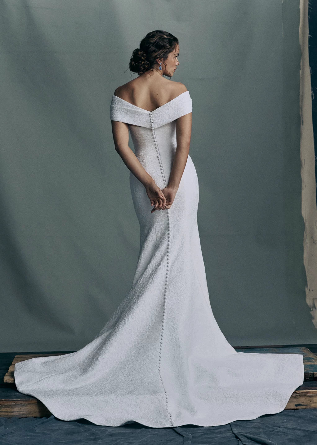 Back view of the Sera gown by Anne Barge is a strapless rounded cat-eye fit and flare gown in Spanish Lacework Jacquard, accented by diagonal draping across the skirt and buttons to the hem in Silk White. The Sera gown includes the Sera Shawl, a detachable off-the-shoulder shawl in Lacework Jacquard.
