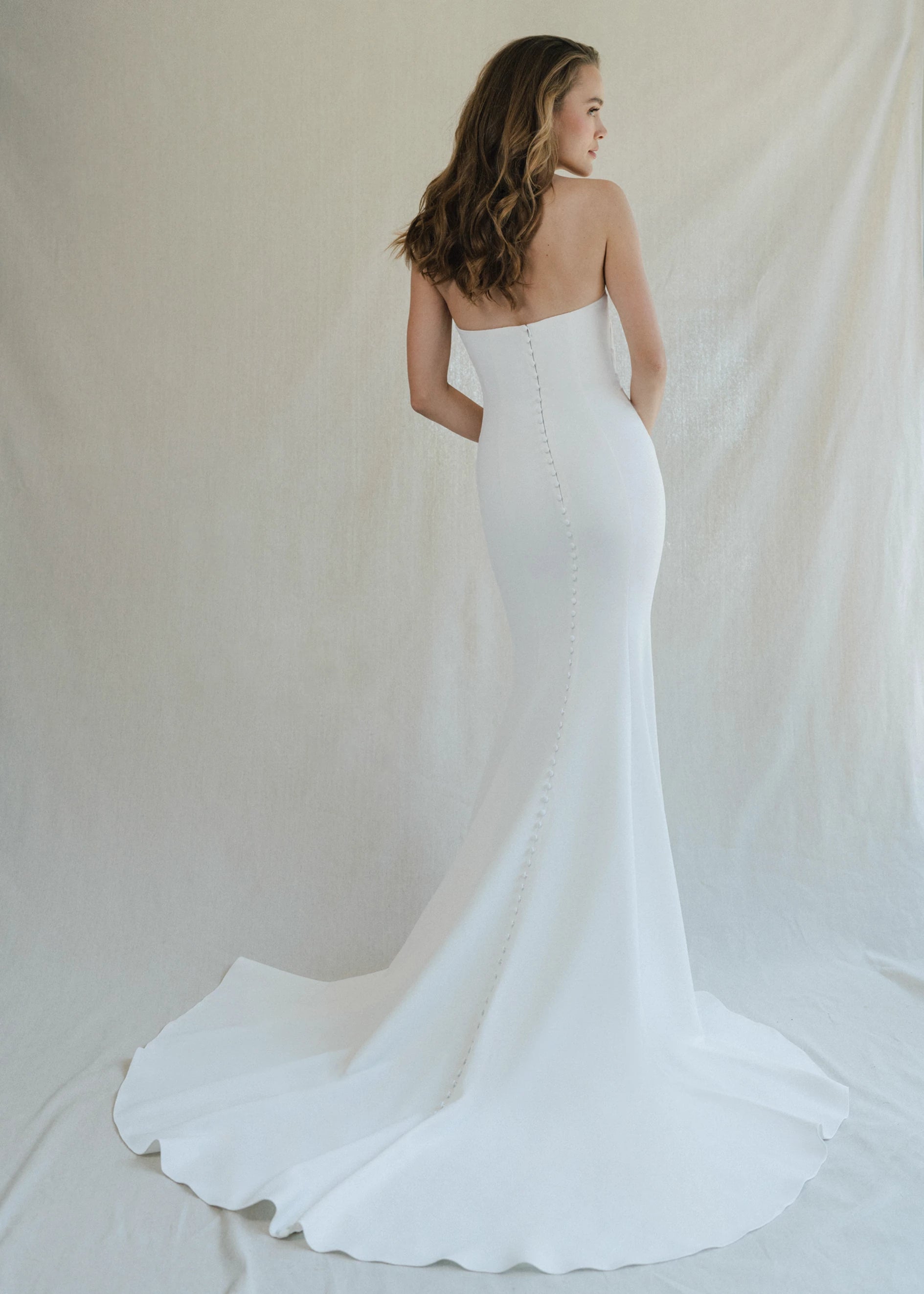The Ryder gown is a strapless V-notch fit and flare gown of Stretch Crepe with figure flattering drape across the front and buttons to the hem in Silk White.