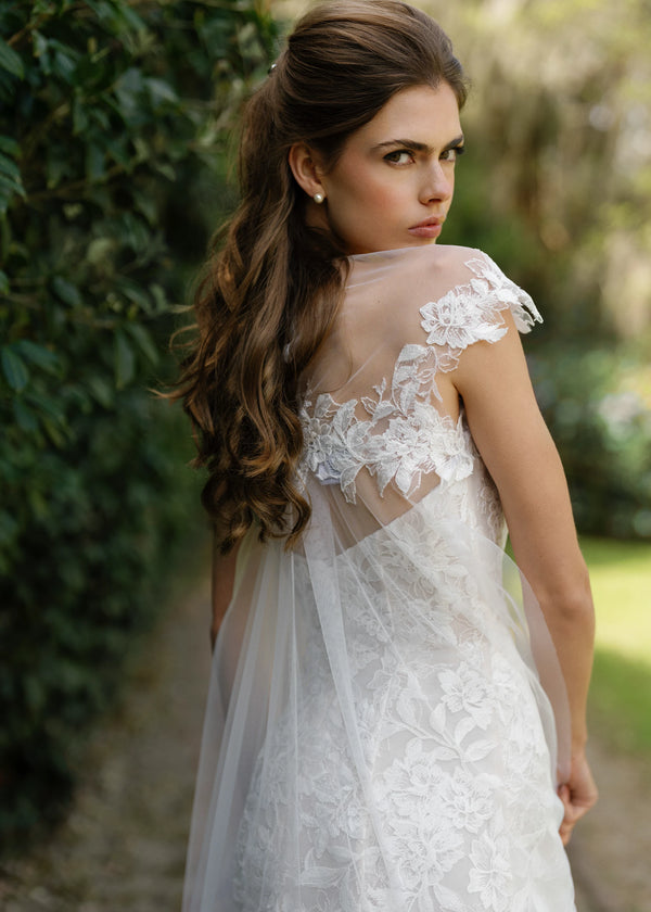 The Cromwell Cape by Anne Barge is an Illusion tulle cape with cascading Threaded Floral Embroidery, attaching at the front neckline and extending into a chapel length train in Silk White. 
