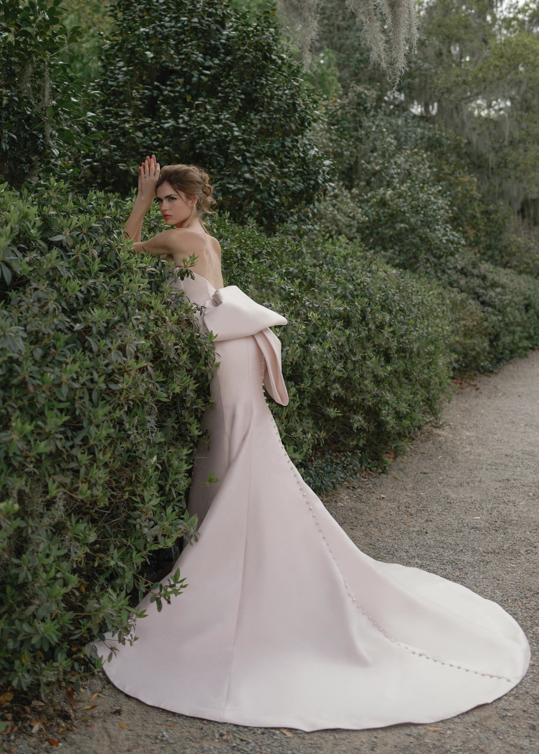 The Daniels gown is a fit and flare gown of Kalika featuring an asymmetrically draped bodice cuff, oversized sculptural bow, and buttons to the hem.