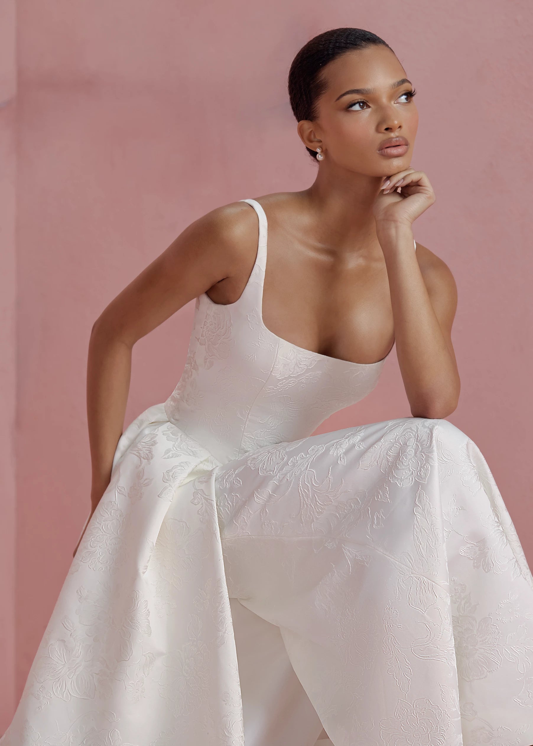 The Elodie gown by Anne Barge is a square neck column gown in Embossed Jacquard with low back and buttons to the hem, featuring box-pleated attached overskirt in Pearl.