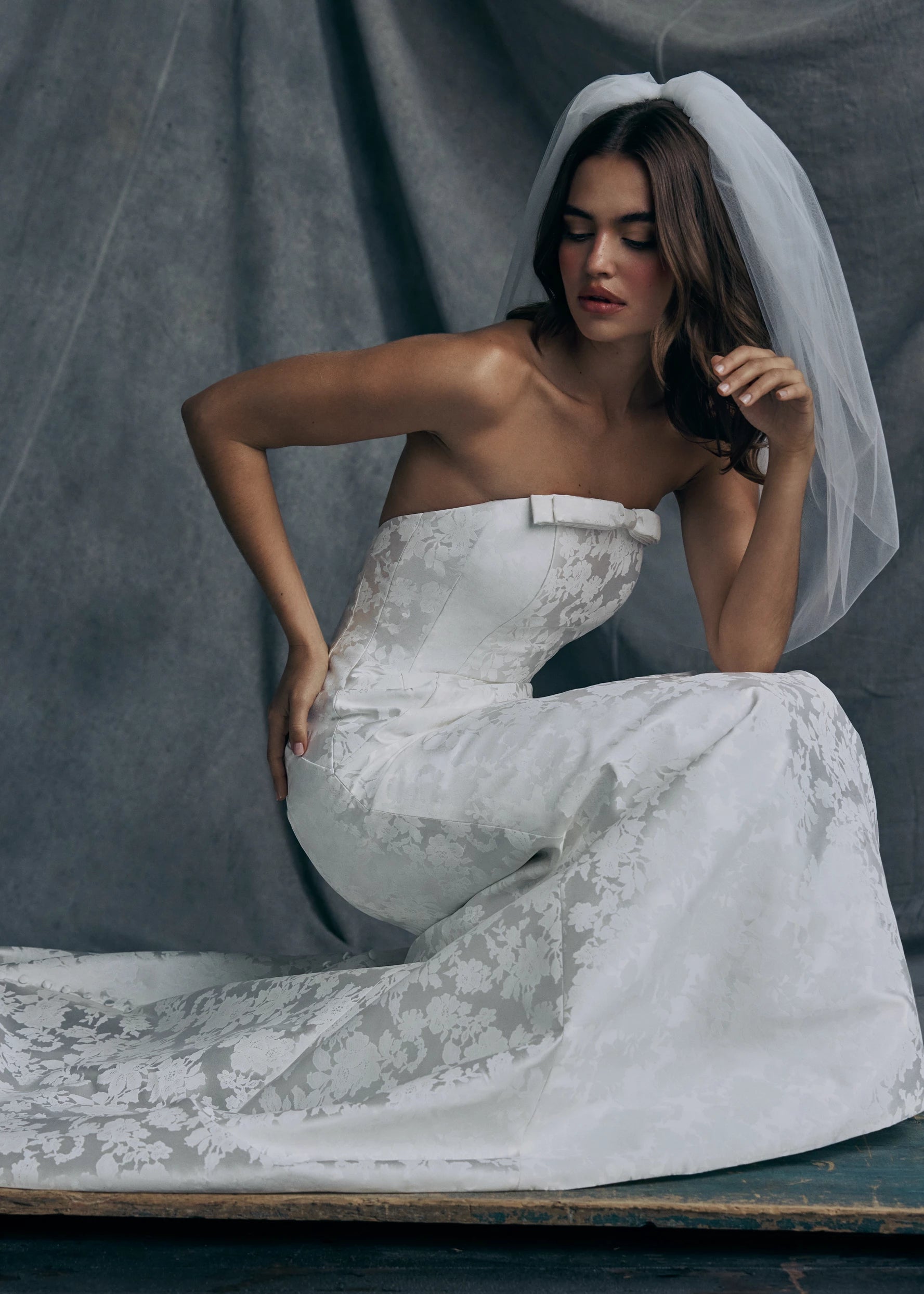 The Elysai gown by Anne Barge is an arched strapless gown in Italian Lace Printed Matte Satin with tailored bow detail, modified A-line skirt and buttons to the hem in Pearl.