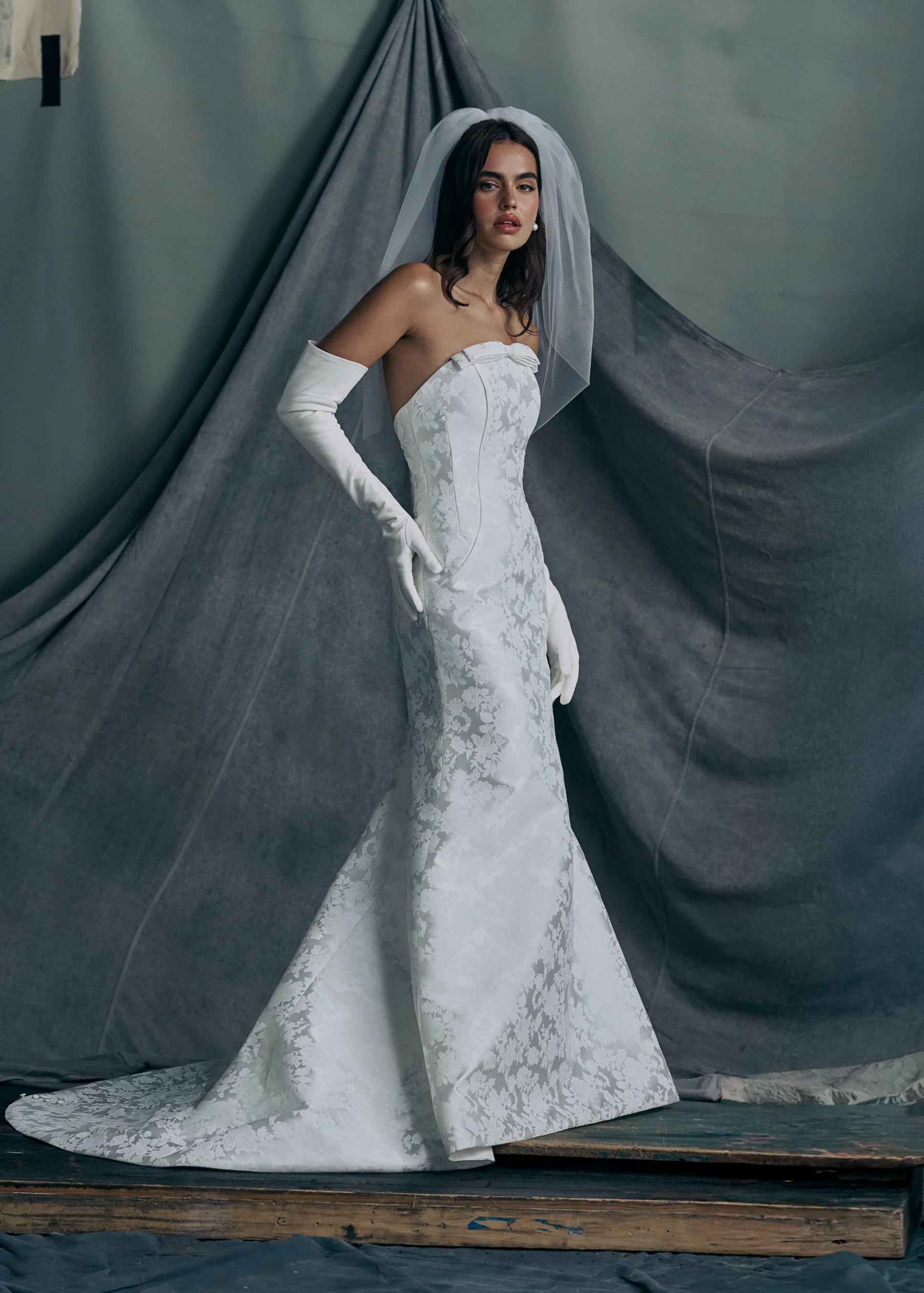 The Elysia gown by Anne Barge is an arched strapless gown in Italian Lace Printed Matte Satin with tailored bow detail, modified A-line skirt and buttons to the hem in Pearl.