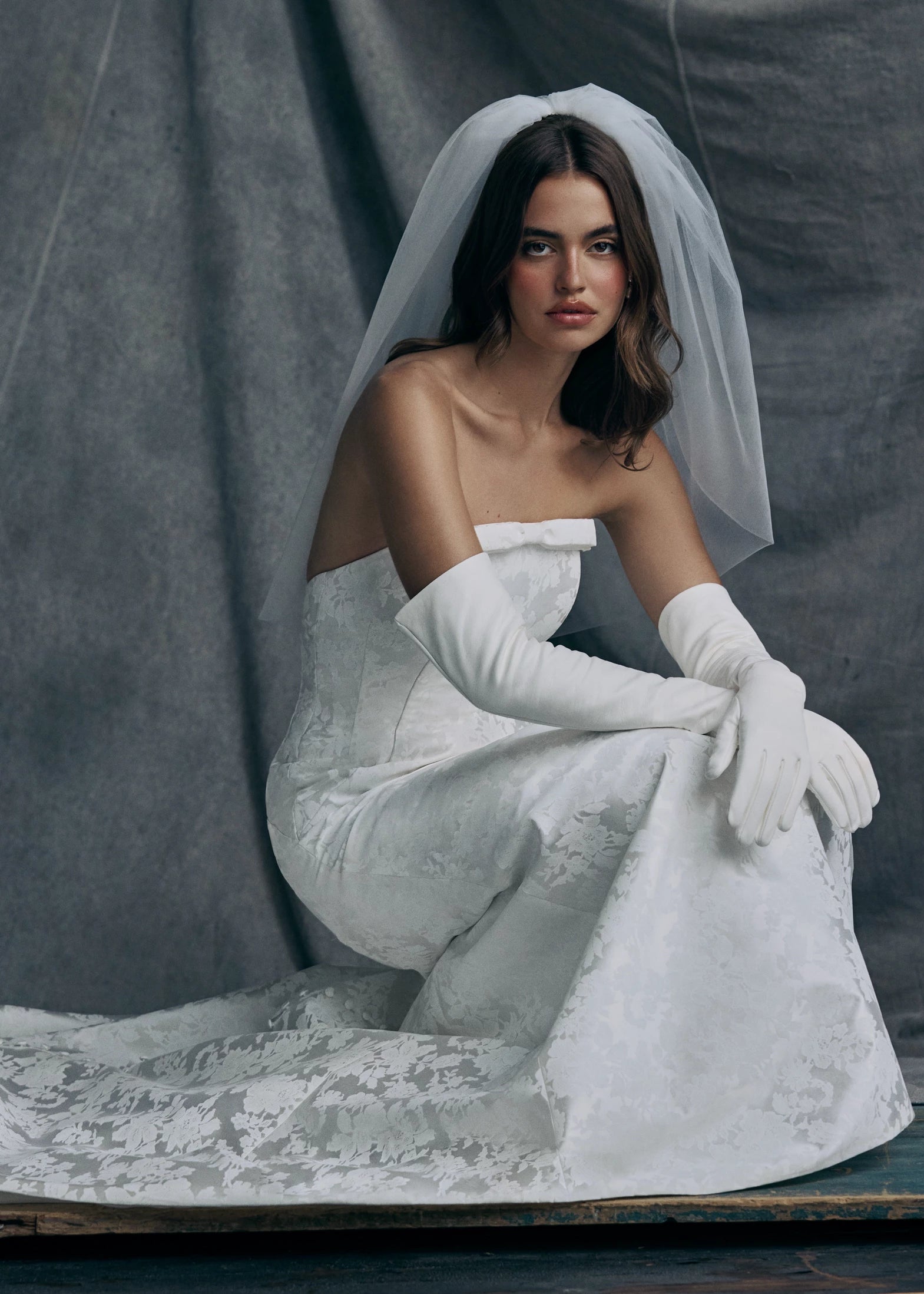 The Elysia gown by Anne Barge is an arched strapless gown in Italian Lace Printed Matte Satin with tailored bow detail, modified A-line skirt and buttons to the hem in Pearl.