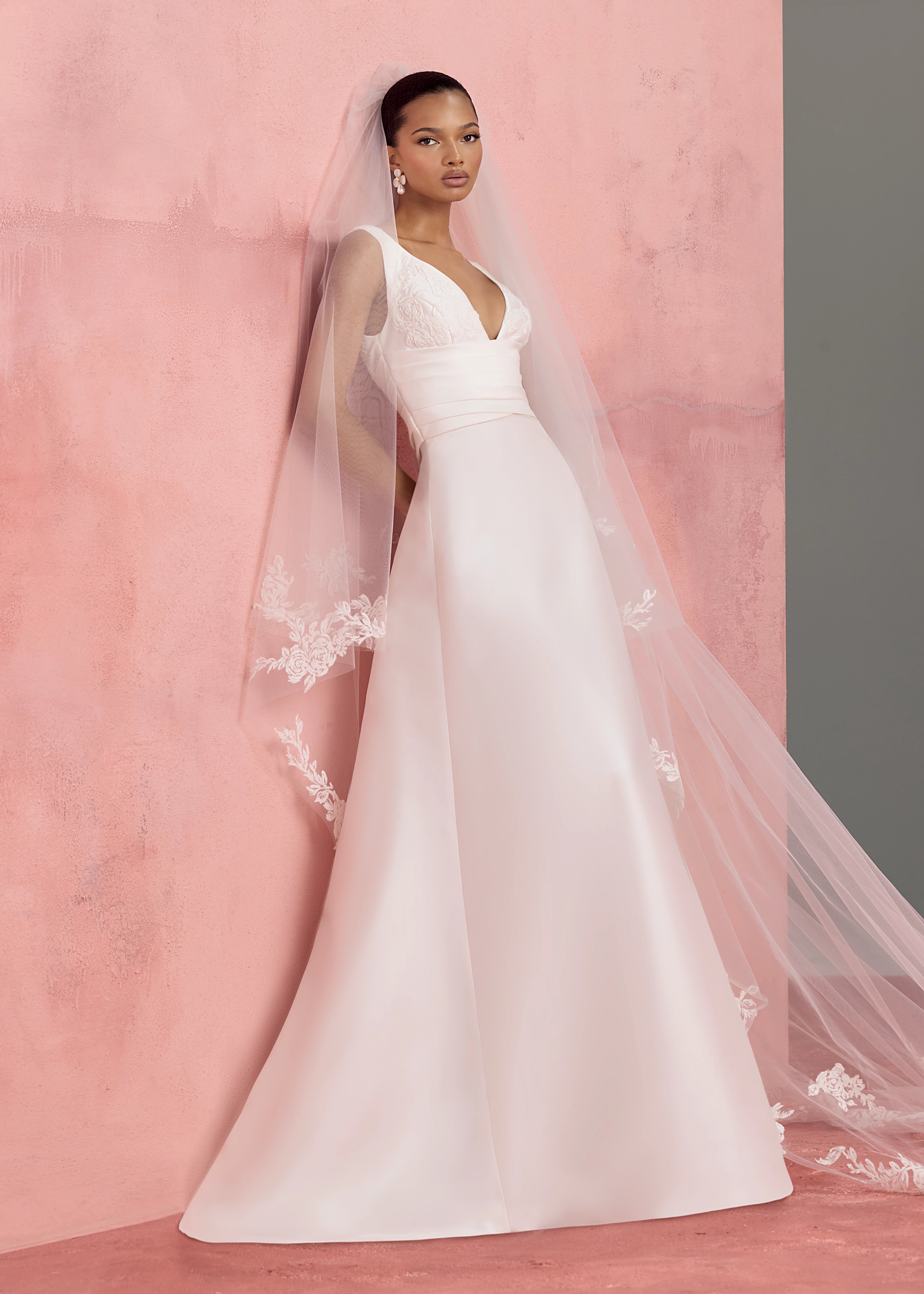 The Joelle Anne Barge gown is a soft Mikado modified A-line gown with plunging V-neckline accented with Thread-Sketched Floral Embroidered Appliques, pleated cummerbund waist, and buttons to the hem in Pearl.
