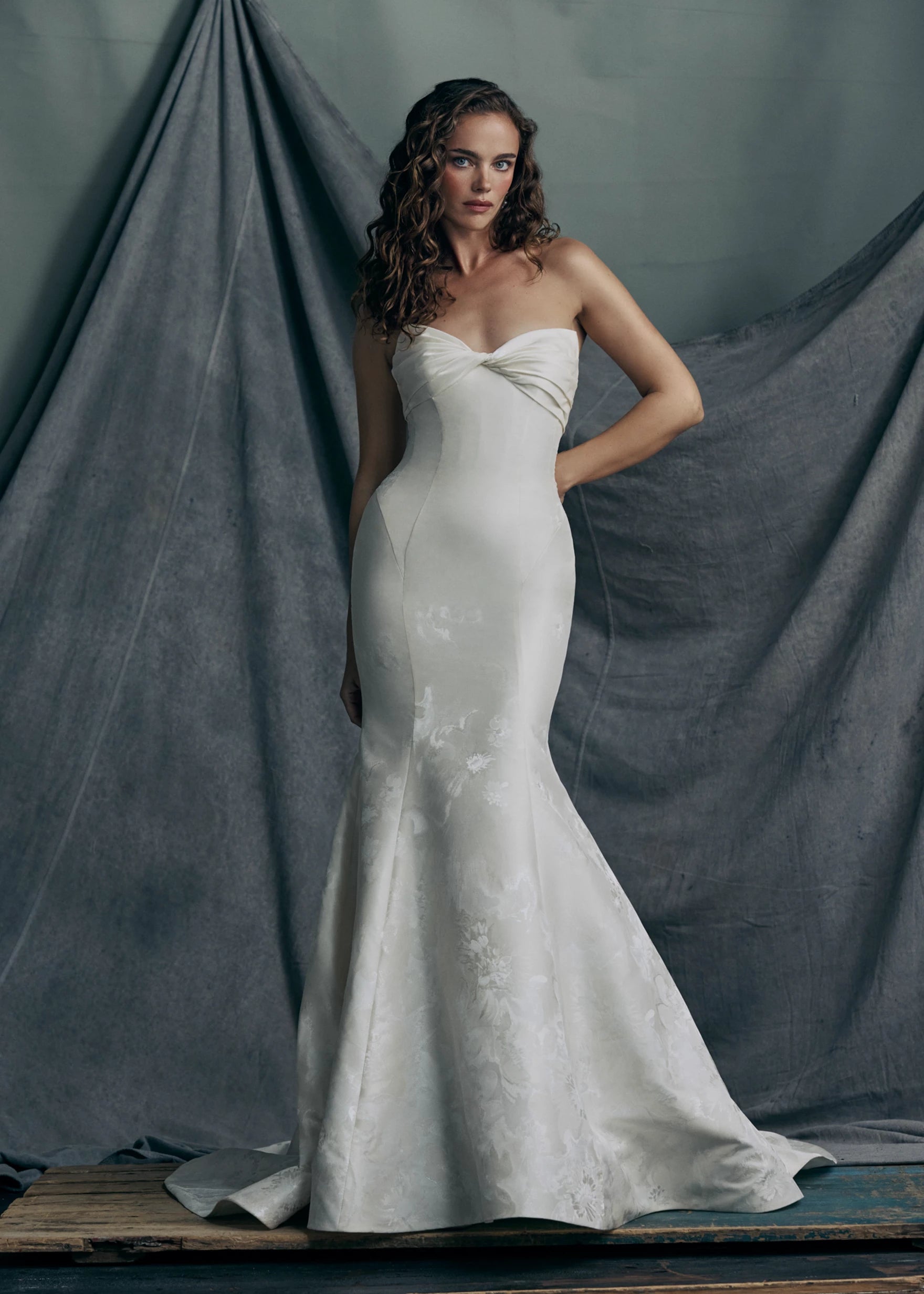 The Justine gown by Anne Barge  is a strapless sweetheart trumpet gown of Italian Floral Matte Satin Jacquard with twisted draped bodice and buttons to the hem in Pearl.