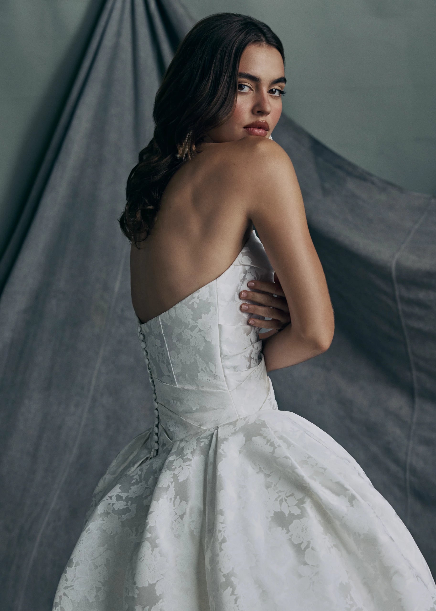 Back view of the Monarch gown by Anne Barge is an arched strapless ballgown in Italian Lace Printed Matte Satin with asymmetrically draped bodice and basque waist on a dramatic box pleated skirt, with buttons to the hem in Pearl.