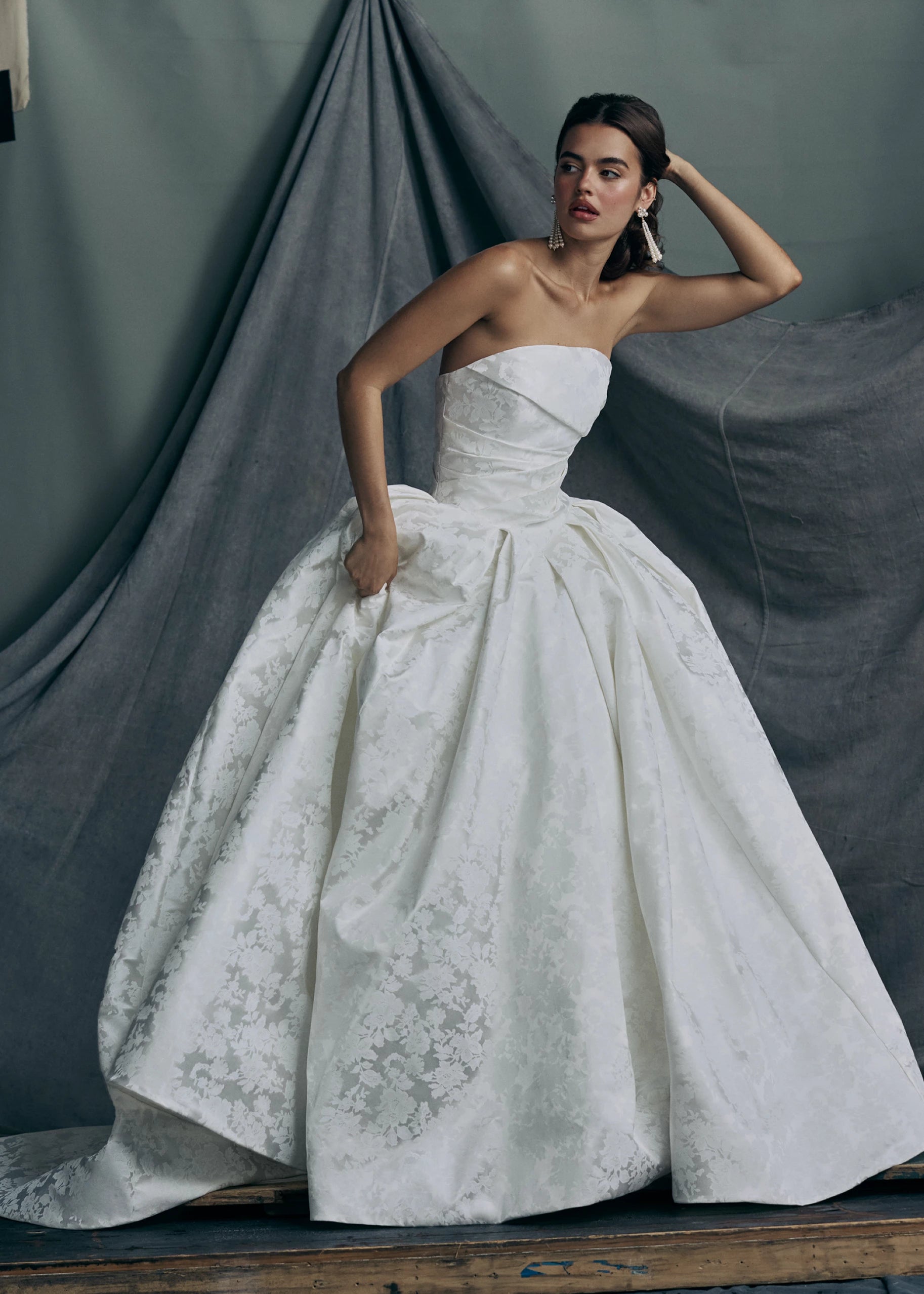 The Monarch gown by Anne Barge is an arched strapless ballgown in Italian Lace Printed Matte Satin with asymmetrically draped bodice and basque waist on a dramatic box pleated skirt, with buttons to the hem in Pearl.