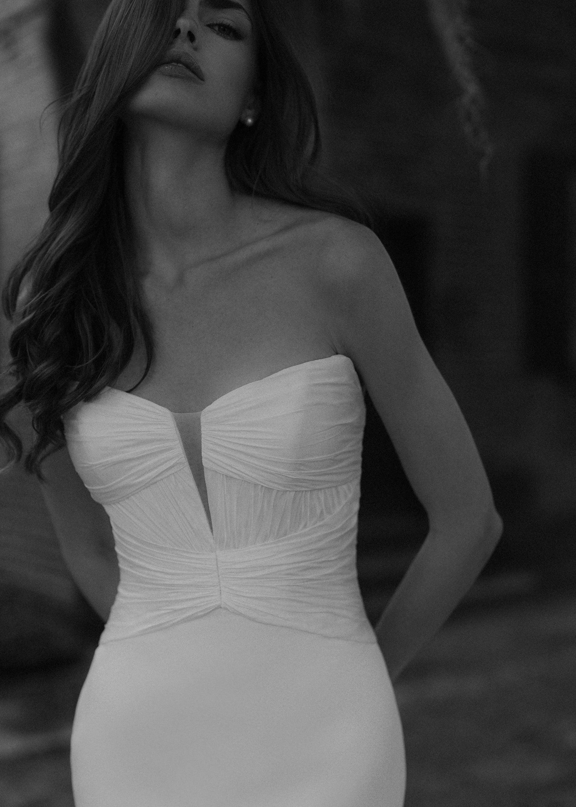 The Montagu gown is a trumpet gown of Italian Kalika with a narrow plunging V-neckline bodice, intricately pleated in Silk Satin Chiffon, and buttons to the top of the godet in Silk White. 