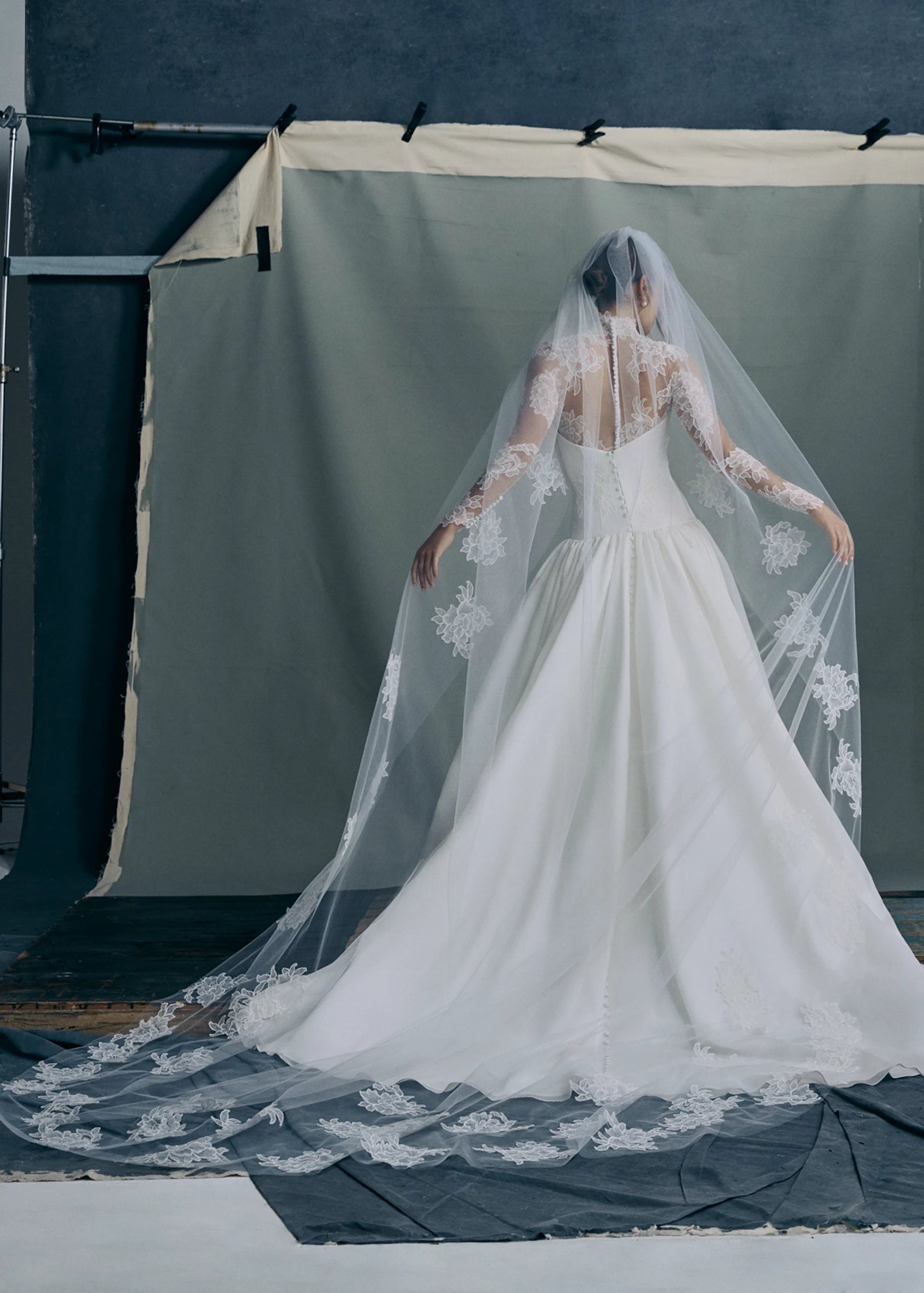 Back view of the Noblesse gown by Anne Barge is a contour strapless drop waist ball gown of Silk Satin Organza with a draped neckline, delicate French Alençon lace appliqués, and a gathered skirt finished with buttons to the hem in Silk White. Paired with the Noblesse Top, an Illusion tulle long sleeve mockneck topper appliquéd with French Alençon lace.