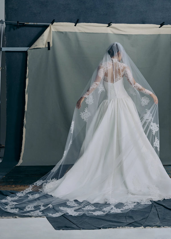 Back view of the Noblesse gown by Anne Barge is a contour strapless drop waist ball gown of Silk Satin Organza with a draped neckline, delicate French Alençon lace appliqués, and a gathered skirt finished with buttons to the hem in Silk White. Paired with the Noblesse Top, an Illusion tulle long sleeve mockneck topper appliquéd with French Alençon lace.