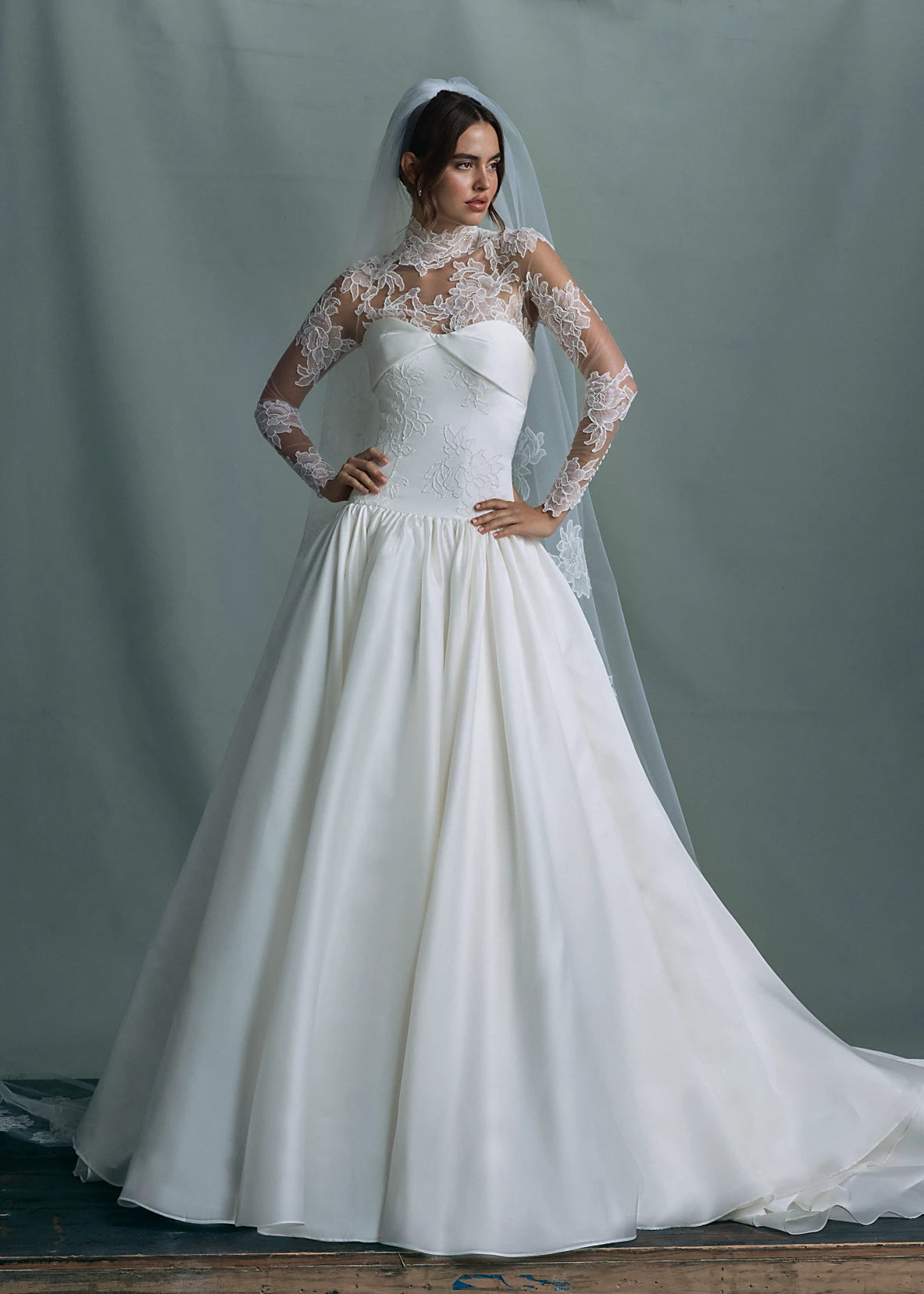 The Noblesse gown by Anne Barge is a contour strapless drop waist ball gown of Silk Satin Organza with a draped neckline, delicate French Alençon lace appliqués, and a gathered skirt finished with buttons to the hem in Silk White.
Paired with the Noblesse Top, an Illusion tulle long sleeve mockneck topper appliquéd with French Alençon lace.