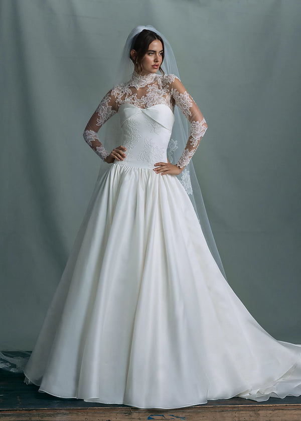 The Noblesse gown by Anne Barge is a contour strapless drop waist ball gown of Silk Satin Organza with a draped neckline, delicate French Alençon lace appliqués, and a gathered skirt finished with buttons to the hem in Silk White.
Paired with the Noblesse Top, an Illusion tulle long sleeve mockneck topper appliquéd with French Alençon lace.