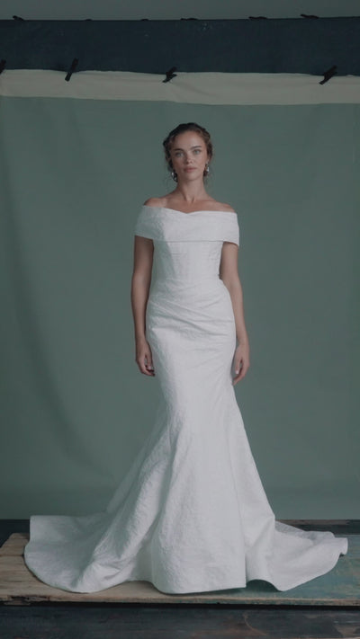 The Sera gown by Anne Barge is a strapless rounded cat-eye fit and flare gown in Spanish Lacework Jacquard, accented by diagonal draping across the skirt and buttons to the hem in Silk White. The Sera gown includes the Sera Shawl, a detachable off-the-shoulder shawl in Lacework Jacquard.