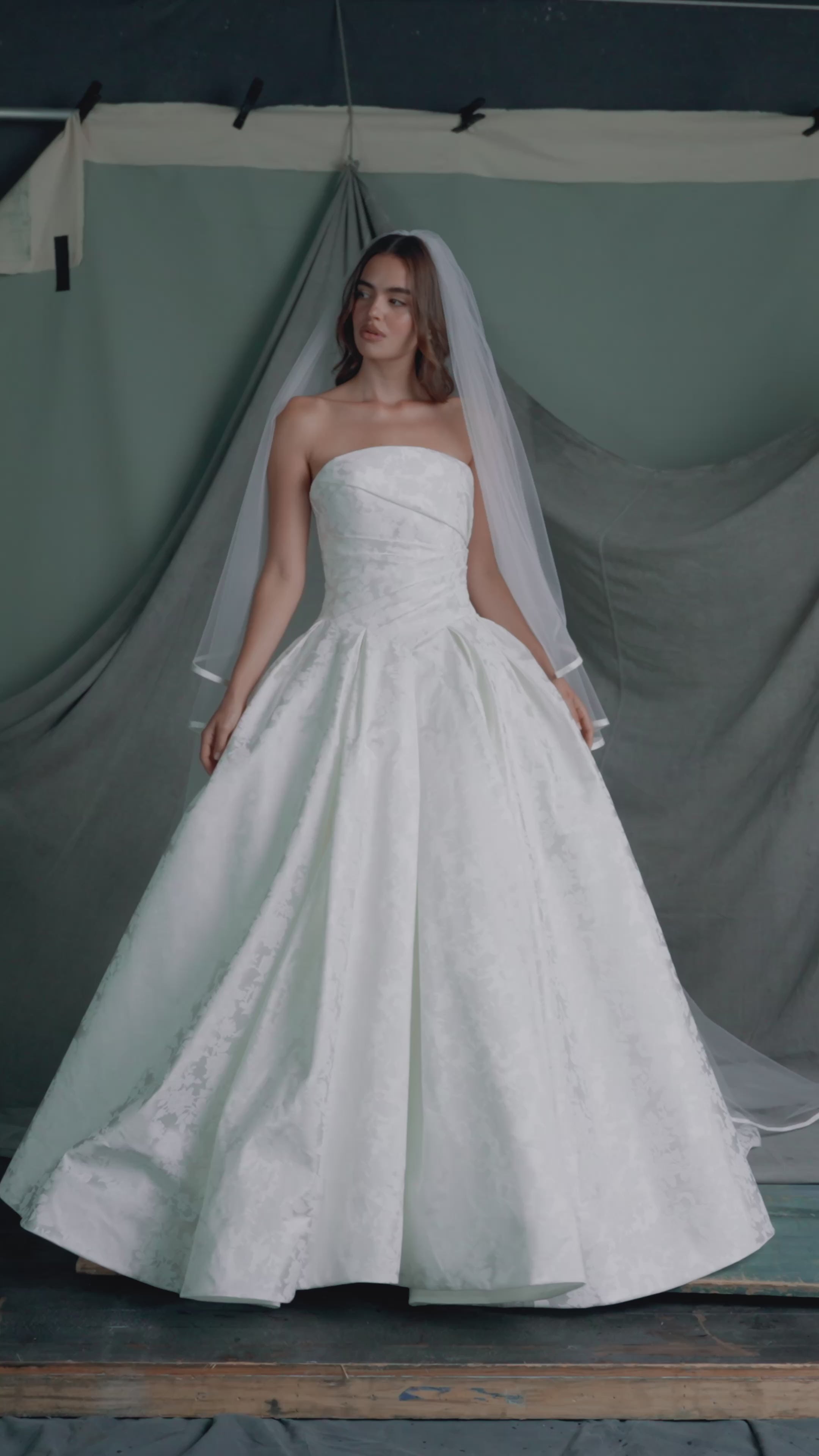 The Monarch gown by Anne Barge is an arched strapless ballgown in Italian Lace Printed Matte Satin with asymmetrically draped bodice and basque waist on a dramatic box pleated skirt, with buttons to the hem in Pearl.
