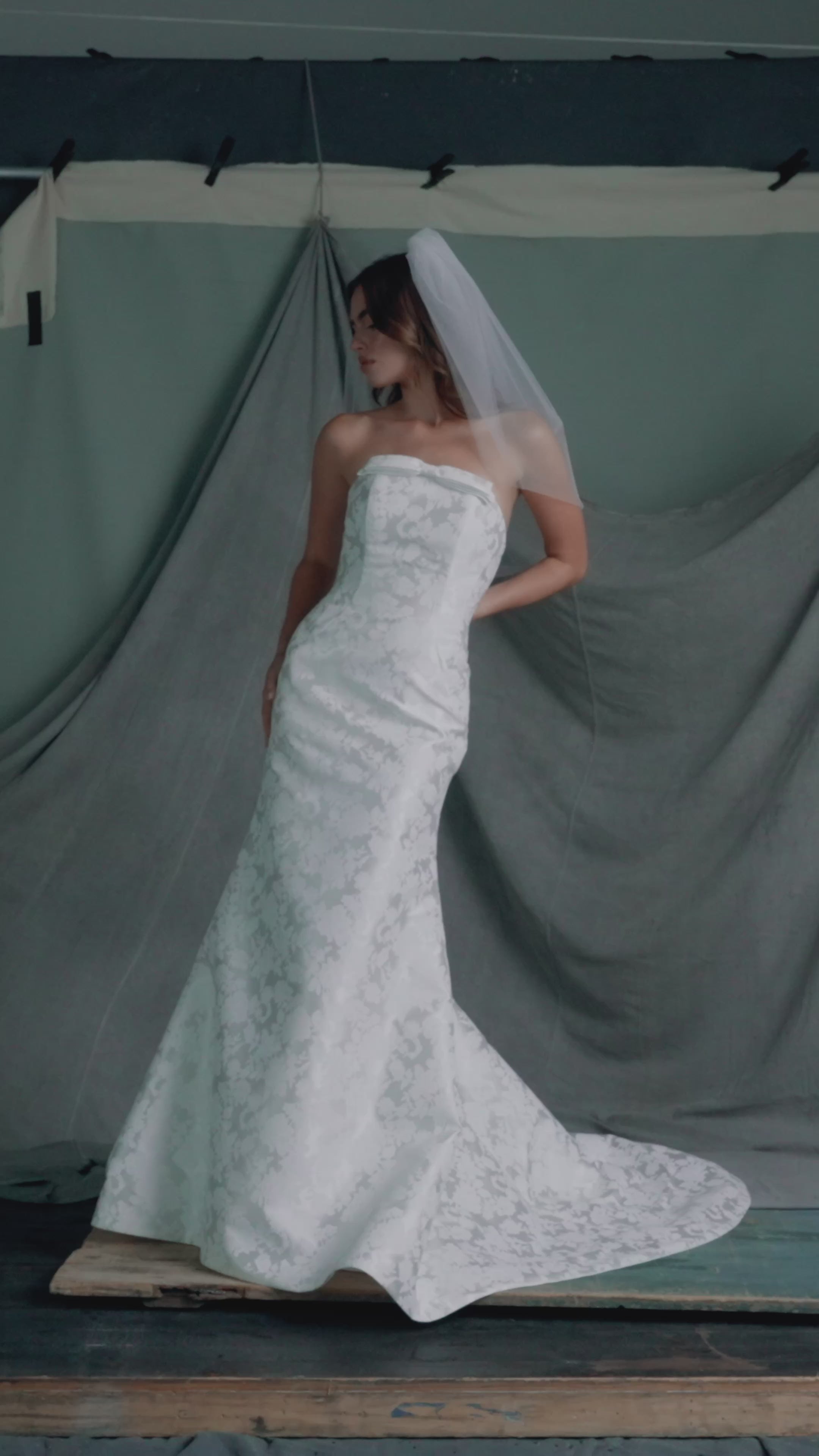 The Elysia gown by Anne Barge is an arched strapless gown in Italian Lace Printed Matte Satin with tailored bow detail, modified A-line skirt and buttons to the hem in Pearl.