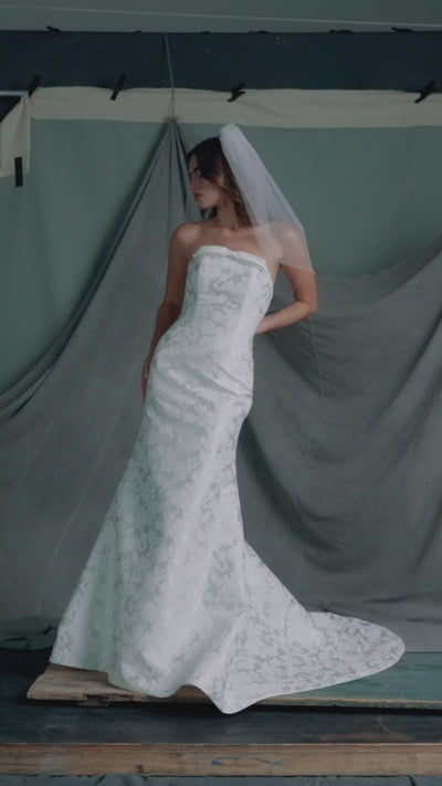 The Elysia gown by Anne Barge is an arched strapless gown in Italian Lace Printed Matte Satin with tailored bow detail, modified A-line skirt and buttons to the hem in Pearl.