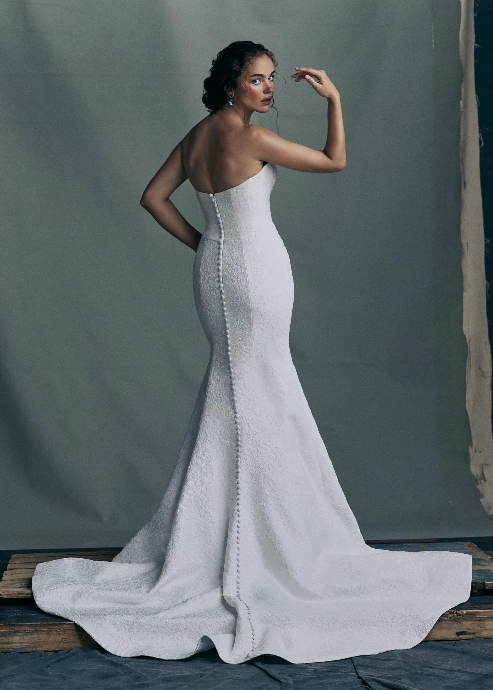 Back view of the Sera gown by Anne Barge is a strapless rounded cat-eye fit and flare gown in Spanish Lacework Jacquard, accented by diagonal draping across the skirt and buttons to the hem in Silk White. The Sera gown includes the Sera Shawl, a detachable off-the-shoulder shawl in Lacework Jacquard.